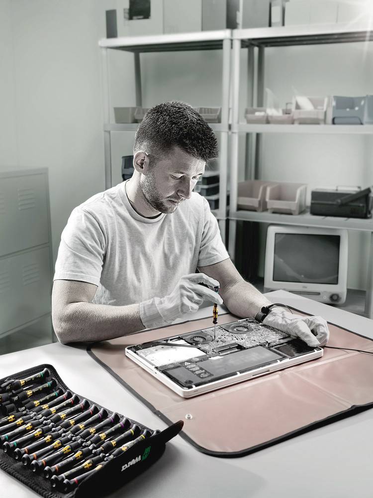 Un homme répare un appareil électronique sur une table. Des outils sont éparpillés autour de lui. Des étagères sont visibles en arrière-plan.