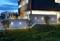 Une maison moderne au crépuscule avec un éclairage extérieur illuminant le mur de béton. Des marches mènent à travers le jardin soigné.