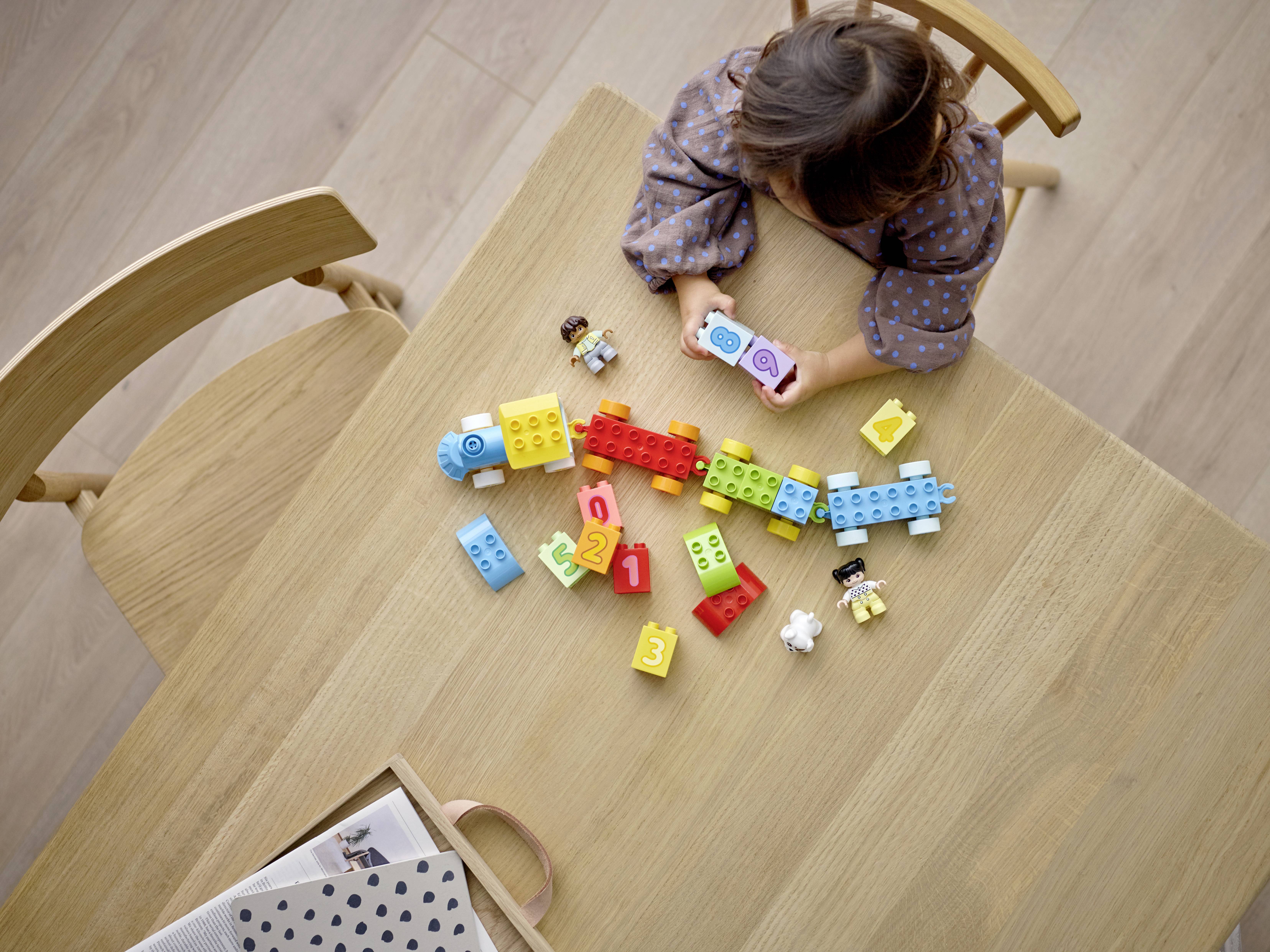 Un enfant joue avec des blocs de construction colorés sur une table en bois, à côté d'une chaise en bois. Les blocs portent des chiffres et des figures, un jeu éducatif.