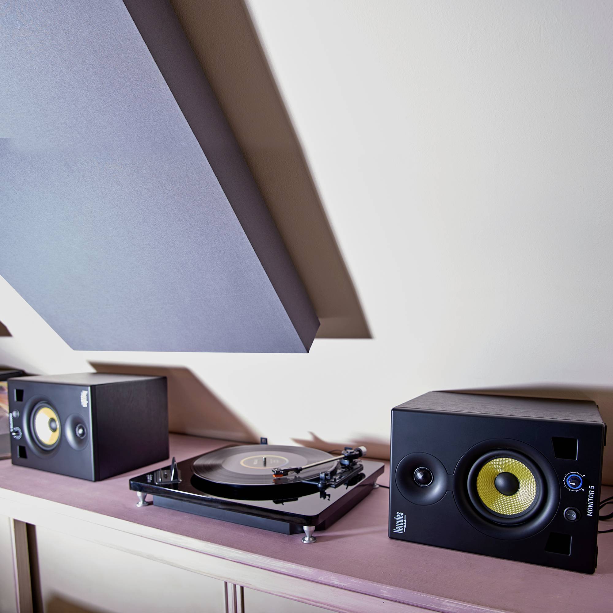 Une platine vinyle est placée entre deux enceintes sur une étagère rose sous un plafond en pente.