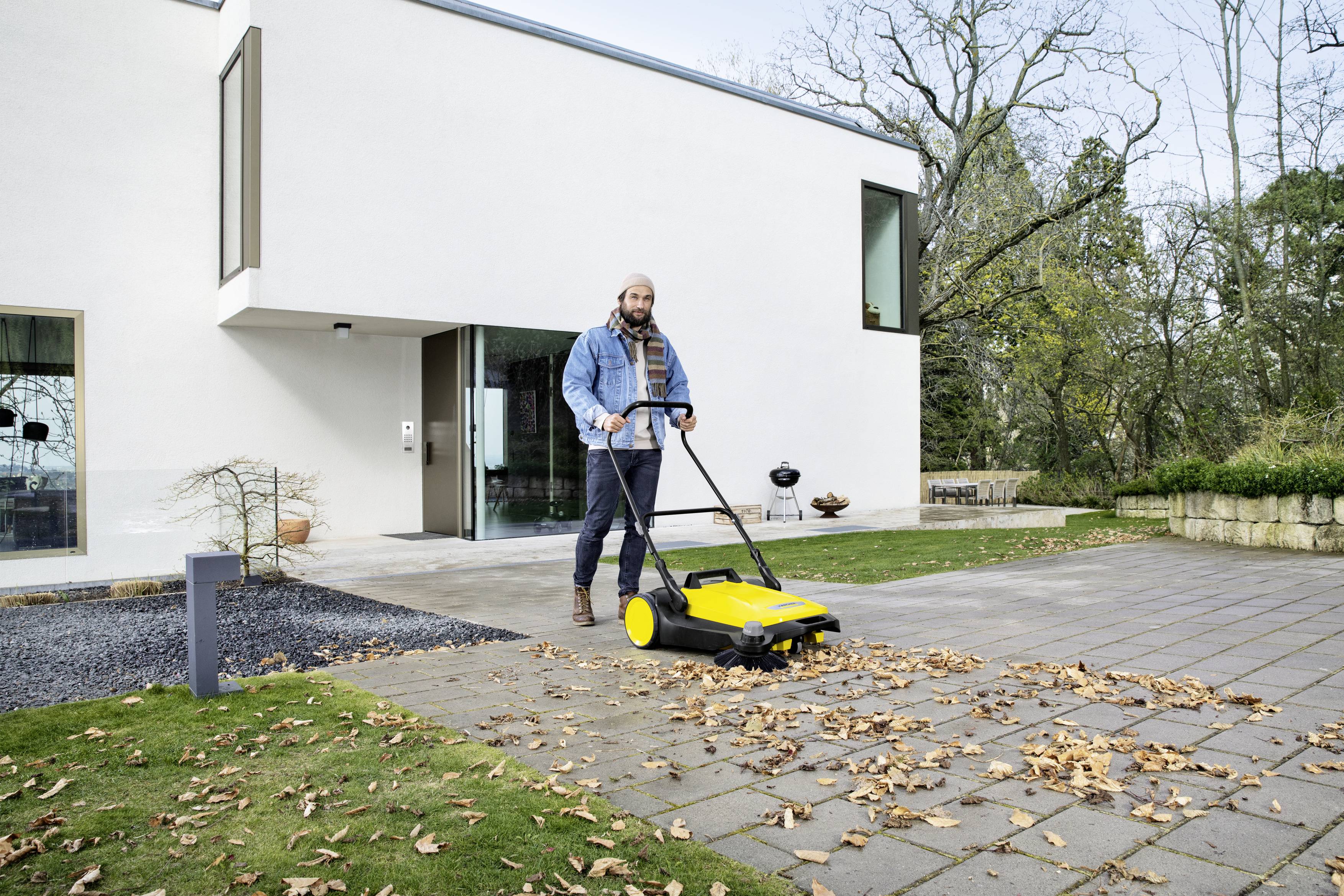 Un homme balaie des feuilles avec un balai de rue jaune pâle sur un chemin pavé devant une maison blanche moderne.