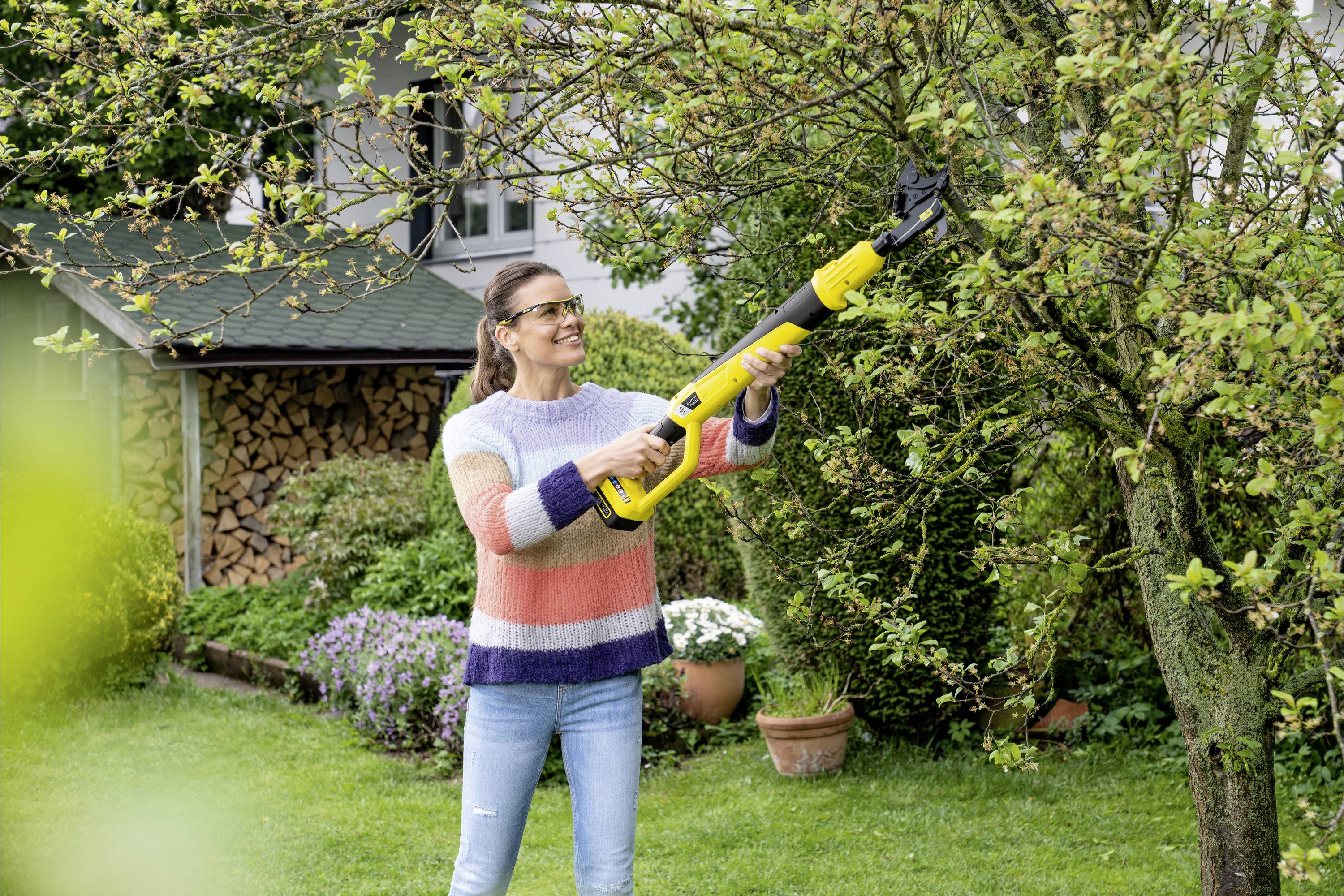 Une femme coupe des branches d'un arbre avec une scie électrique jaune. Elle porte un gilet tricoté coloré et des lunettes de protection dans le jardin.