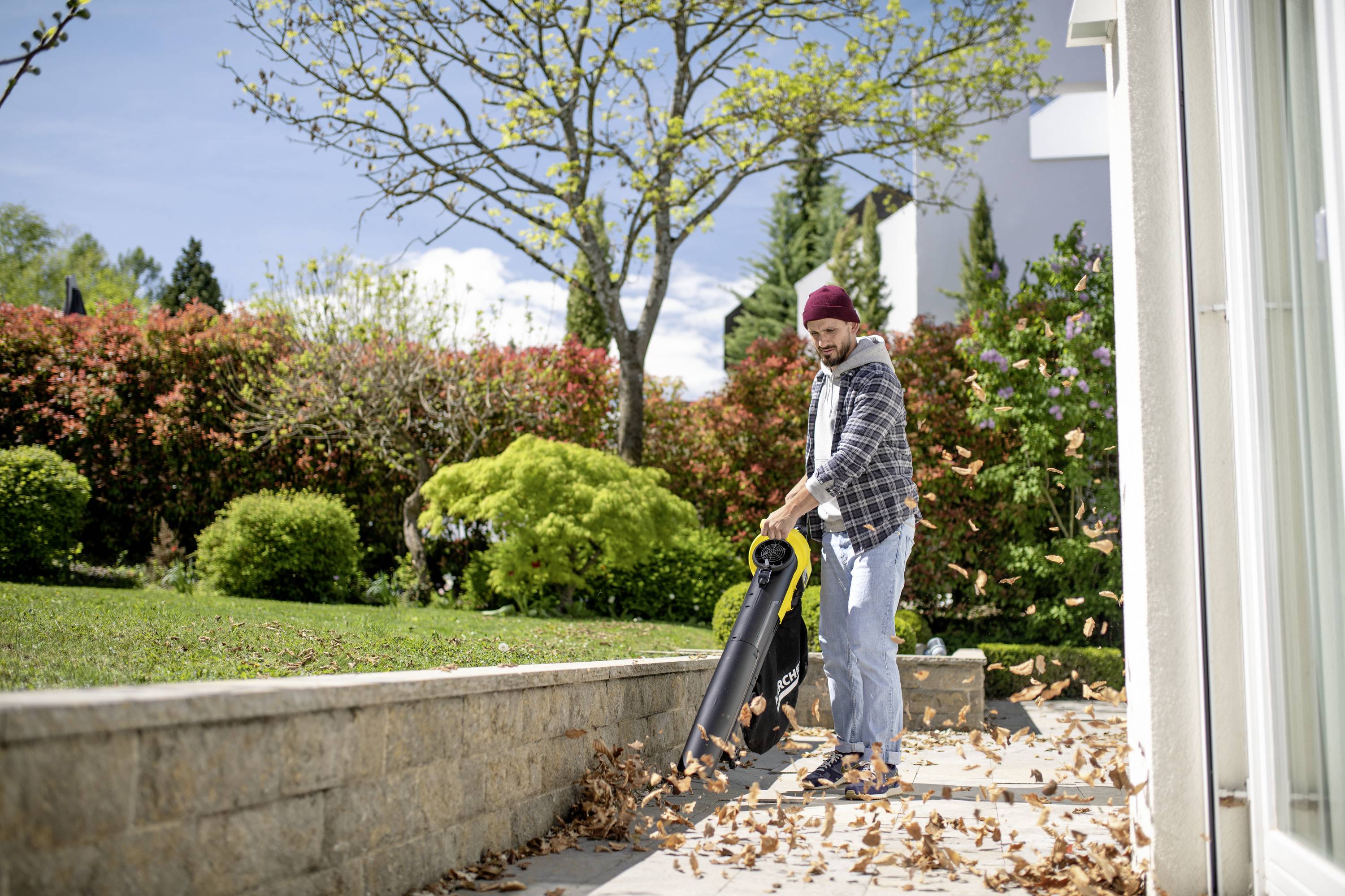 Un homme en tenue décontractée aspire des feuilles d'automne avec un souffleur de feuilles sur une terrasse. À l'arrière-plan, on aperçoit un jardin avec des arbres.