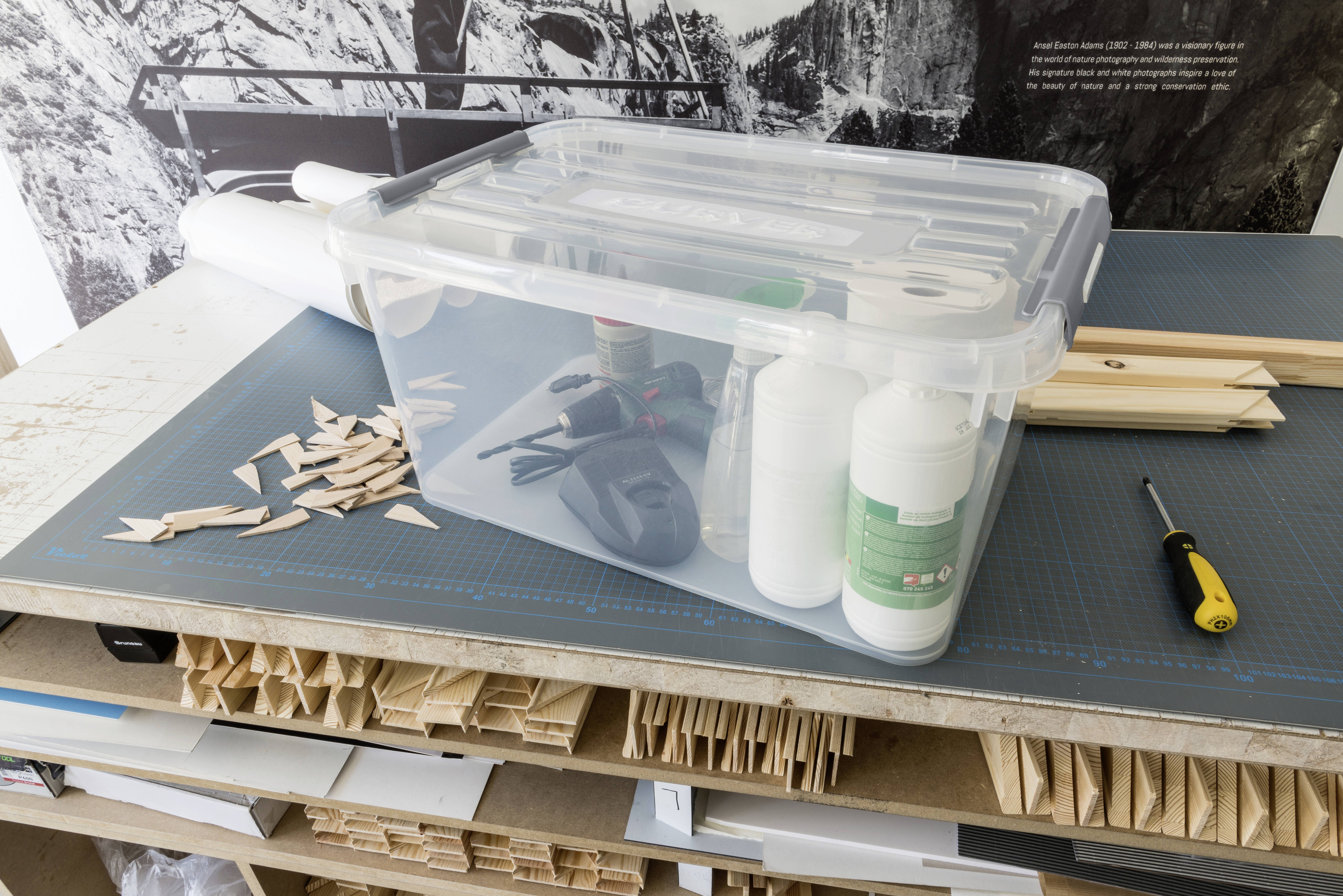 Une boîte en plastique transparent contenant des outils et des matériaux est posée sur une table de travail avec des morceaux de bois et un tournevis.