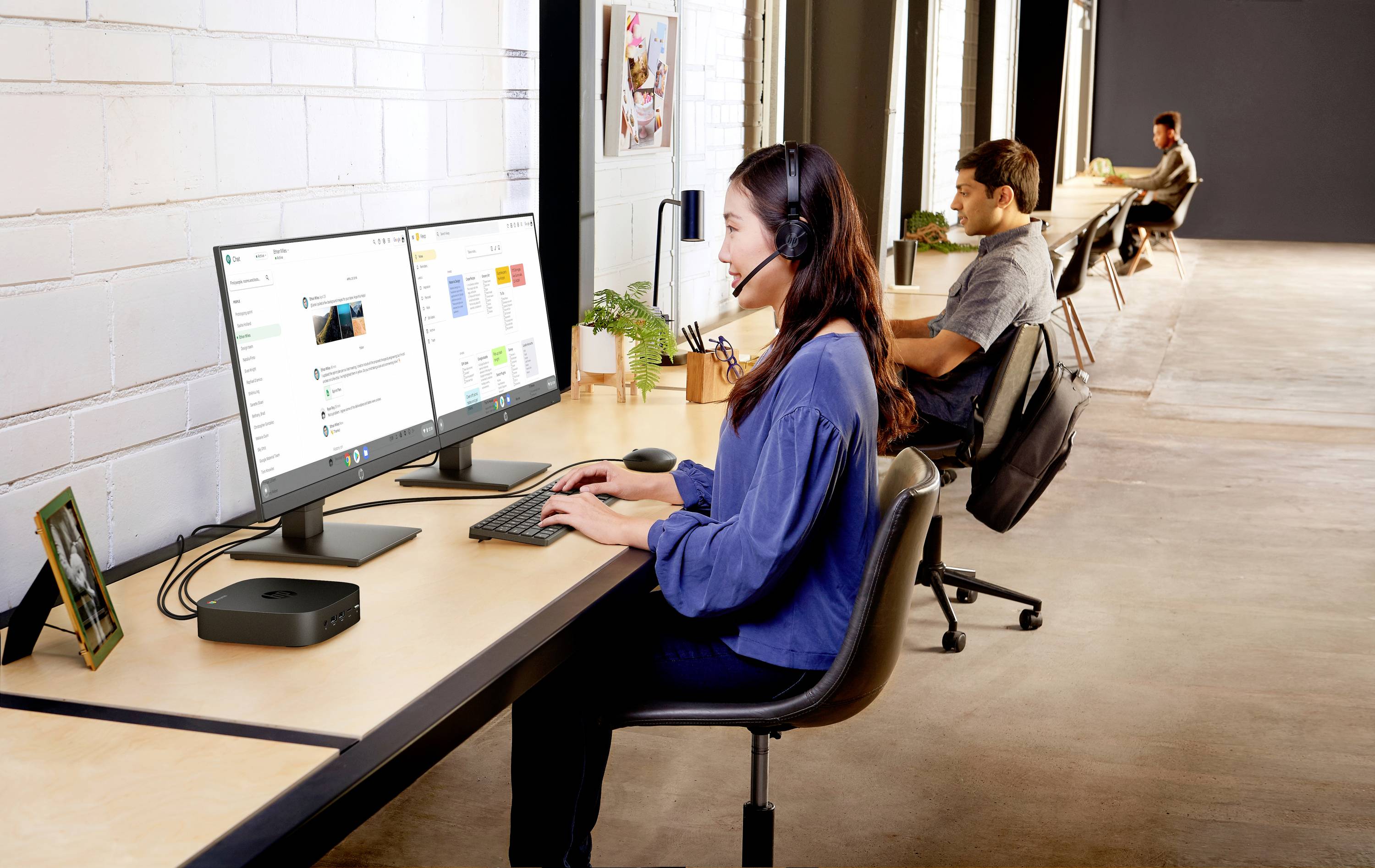 Femme portant un casque de travail, travaillant sur deux écrans dans un bureau moderne ; à l'arrière-plan, un homme travaille sur un ordinateur portable.