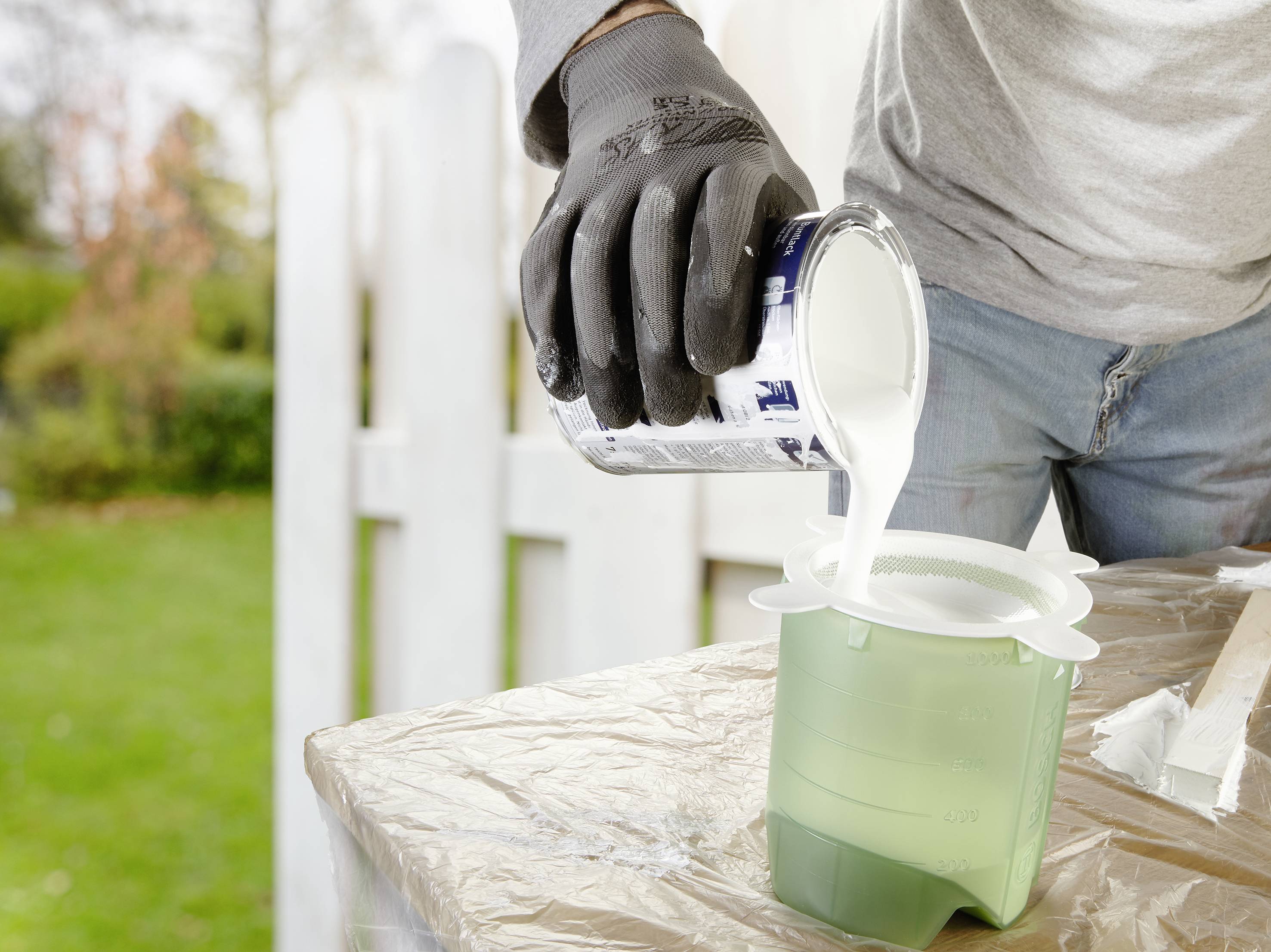 Une personne verse de la peinture blanche d'un bidon dans un récipient vert. La personne porte des gants gris. À l'arrière-plan, on aperçoit un jardin.