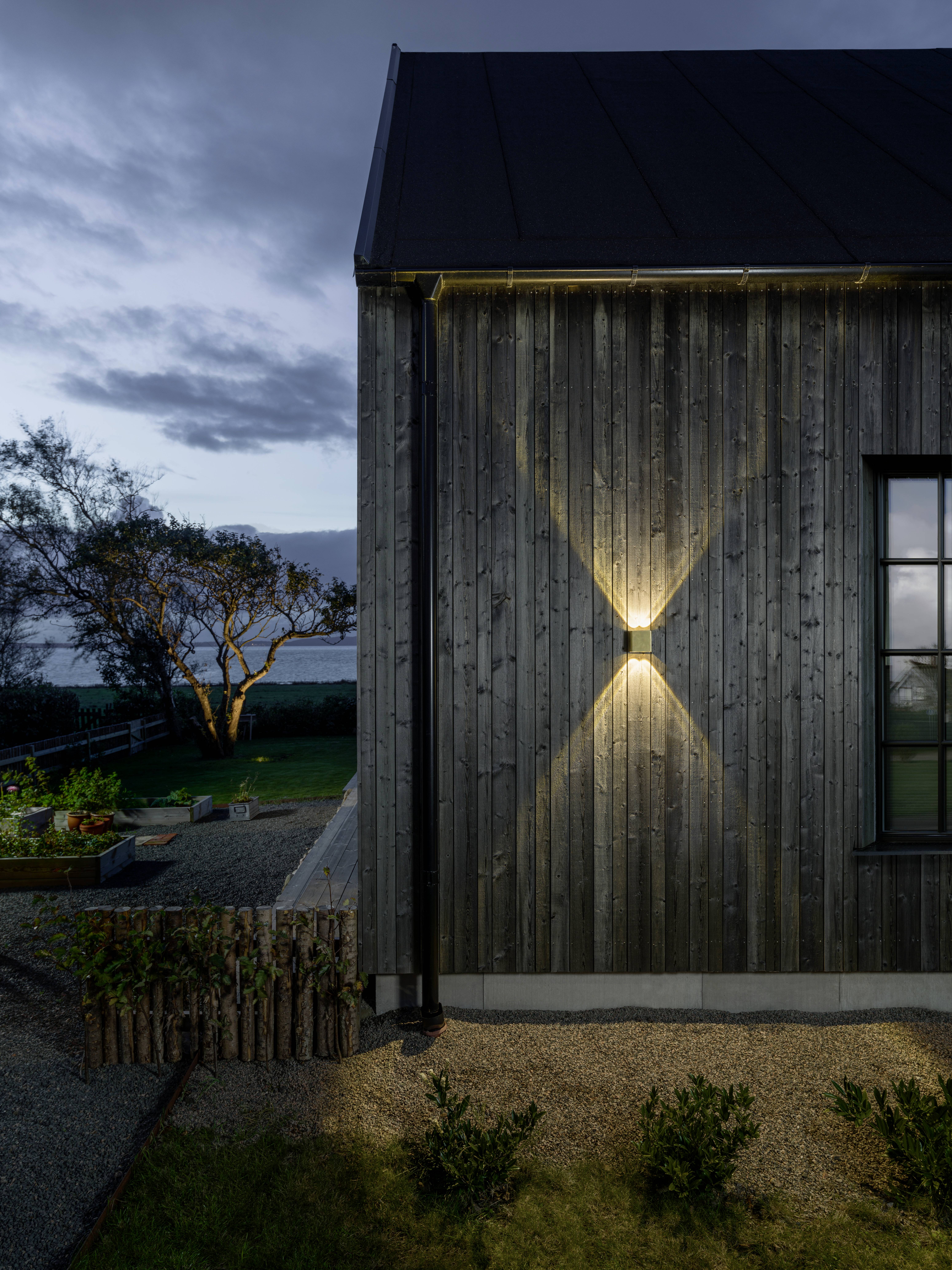Maison recouverte de bois au crépuscule avec un luminaire extérieur allumé projetant un motif en X sur le mur. Arbre et jardin en arrière-plan.