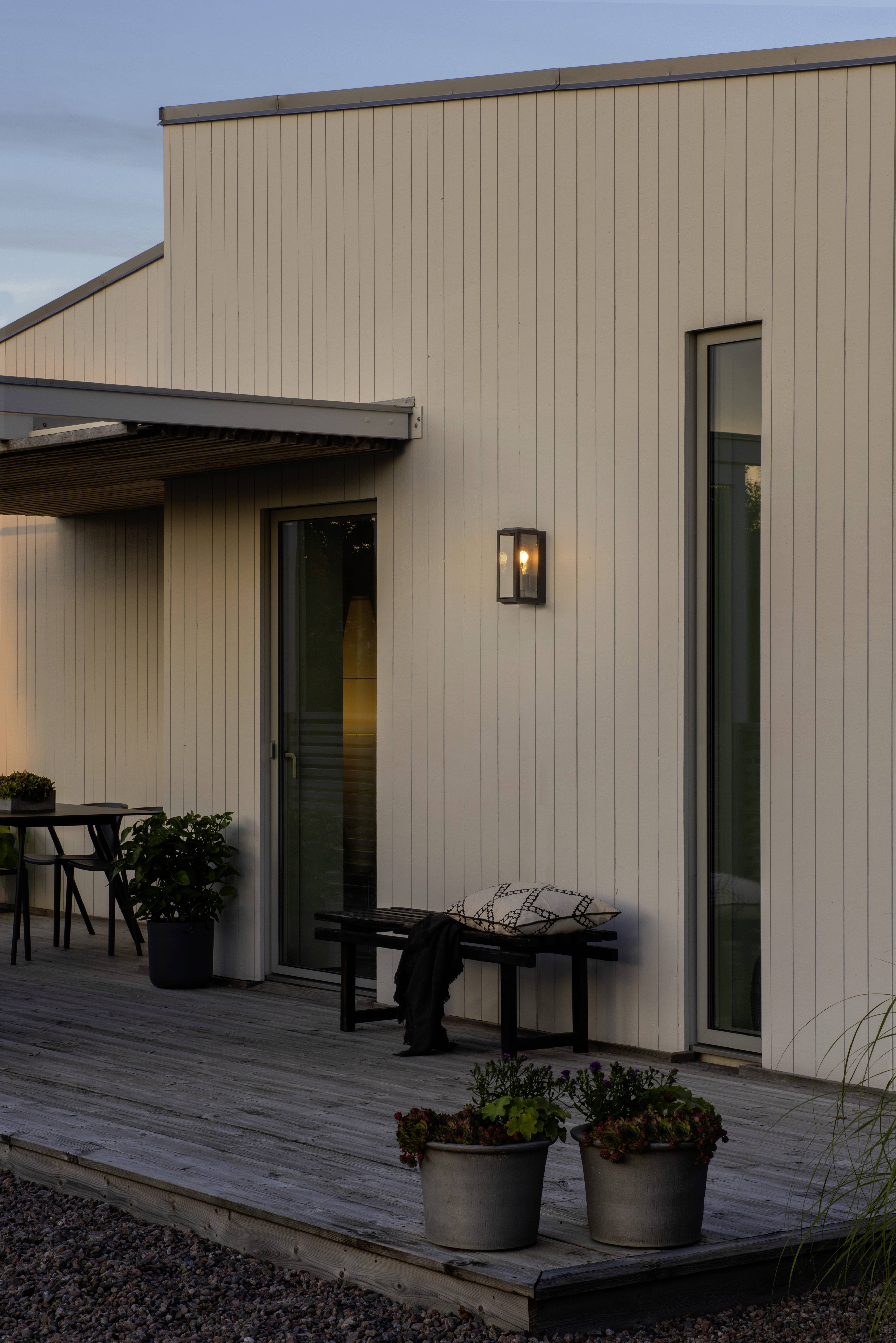 Une maison moderne avec façade en bois au crépuscule. Sur la terrasse en bois se trouvent une table et des chaises. Une lampe brille près de la porte.
