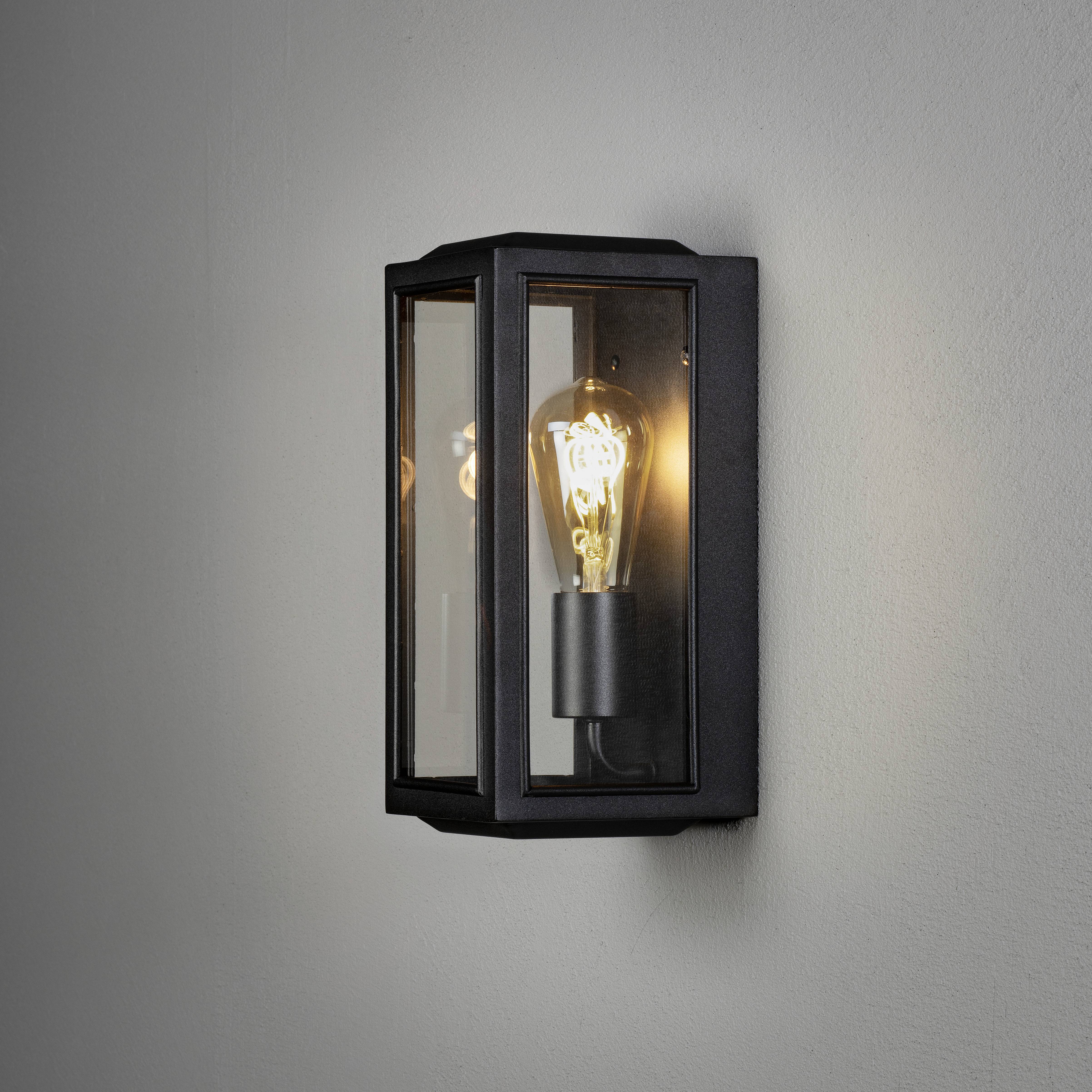 Un applique murale moderne avec un boîtier en verre transparent, éclairant une unique ampoule à lumière chaude sur un mur gris.