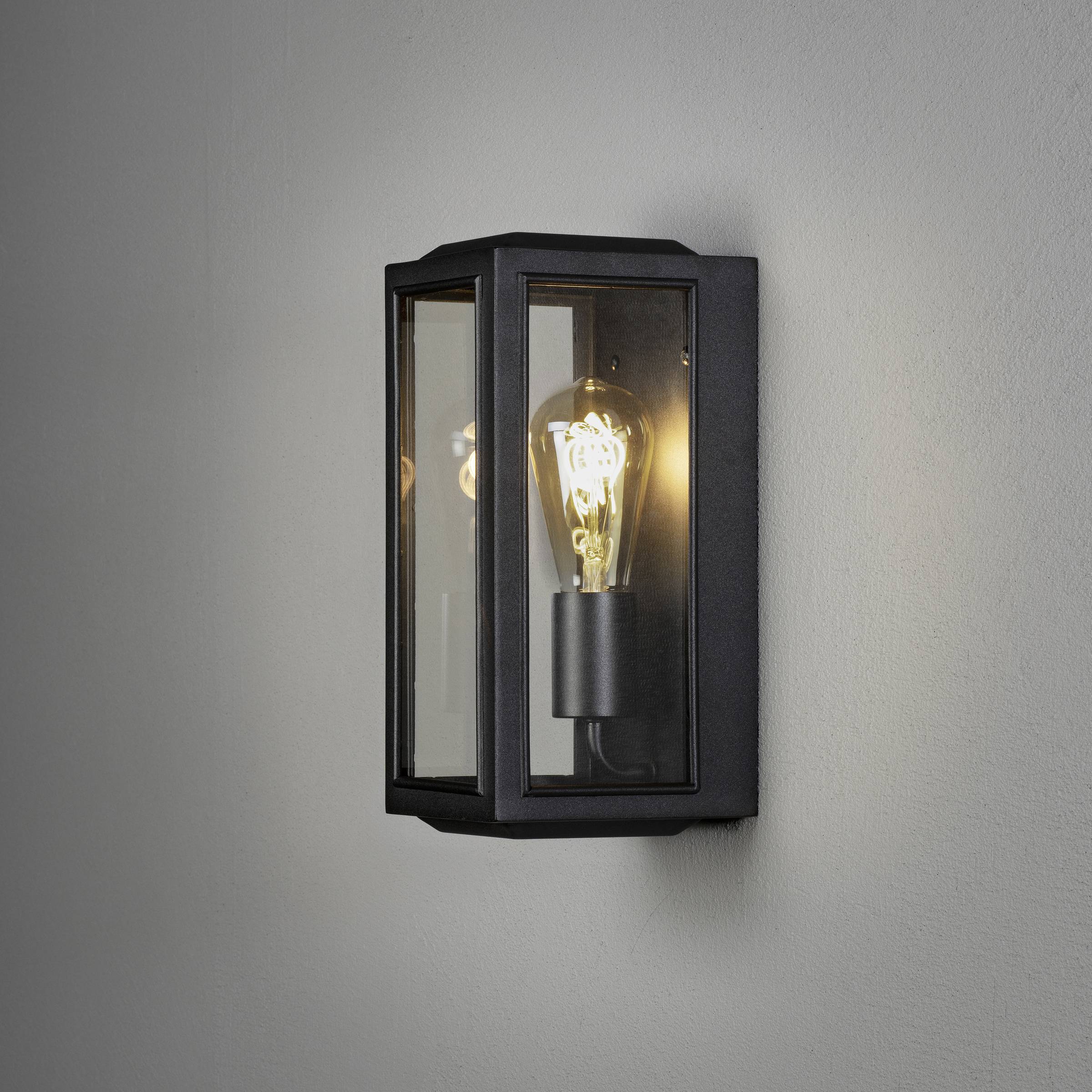 Applique murale noire avec ampoule à l'intérieur d'un boîtier rectangulaire transparent, fixée à un mur gris. Allumée, de style moderne.