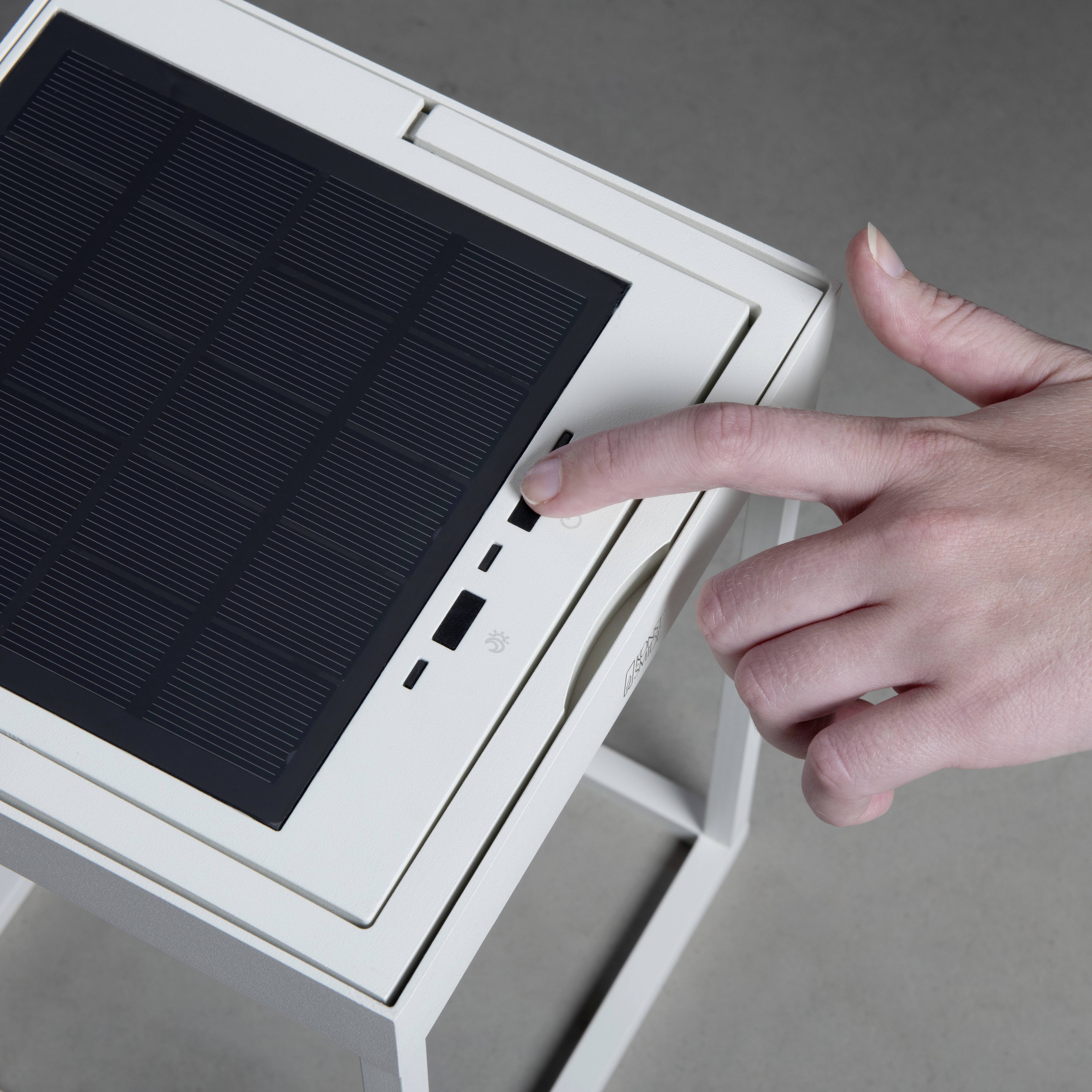 Une main pointe vers un petit panneau solaire carré sur un appareil blanc posé sur une table.
