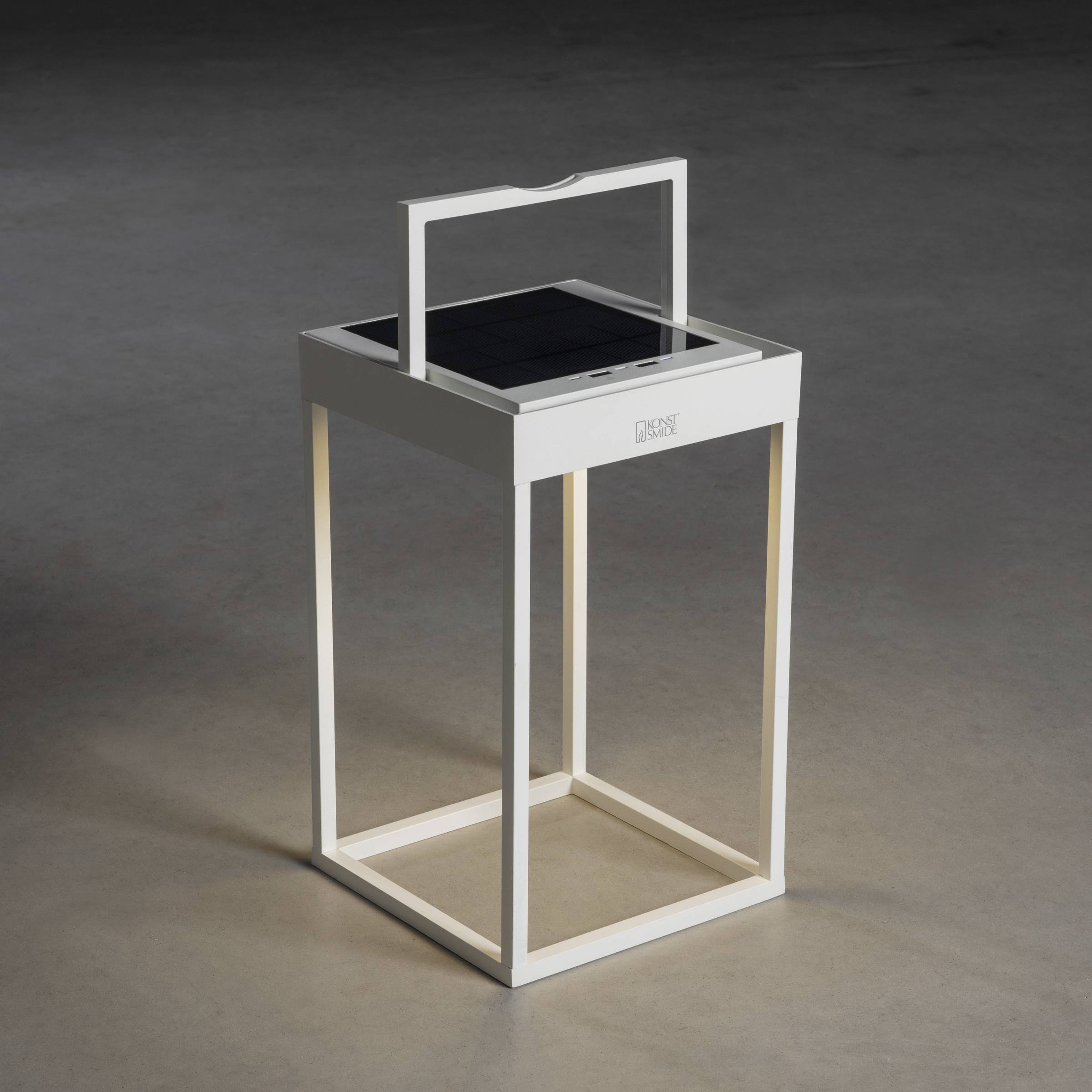 Une lanterne blanche carrée avec un panneau solaire sur le dessus, posée sur un sol gris, diffuse une lumière chaleureuse vers le bas.