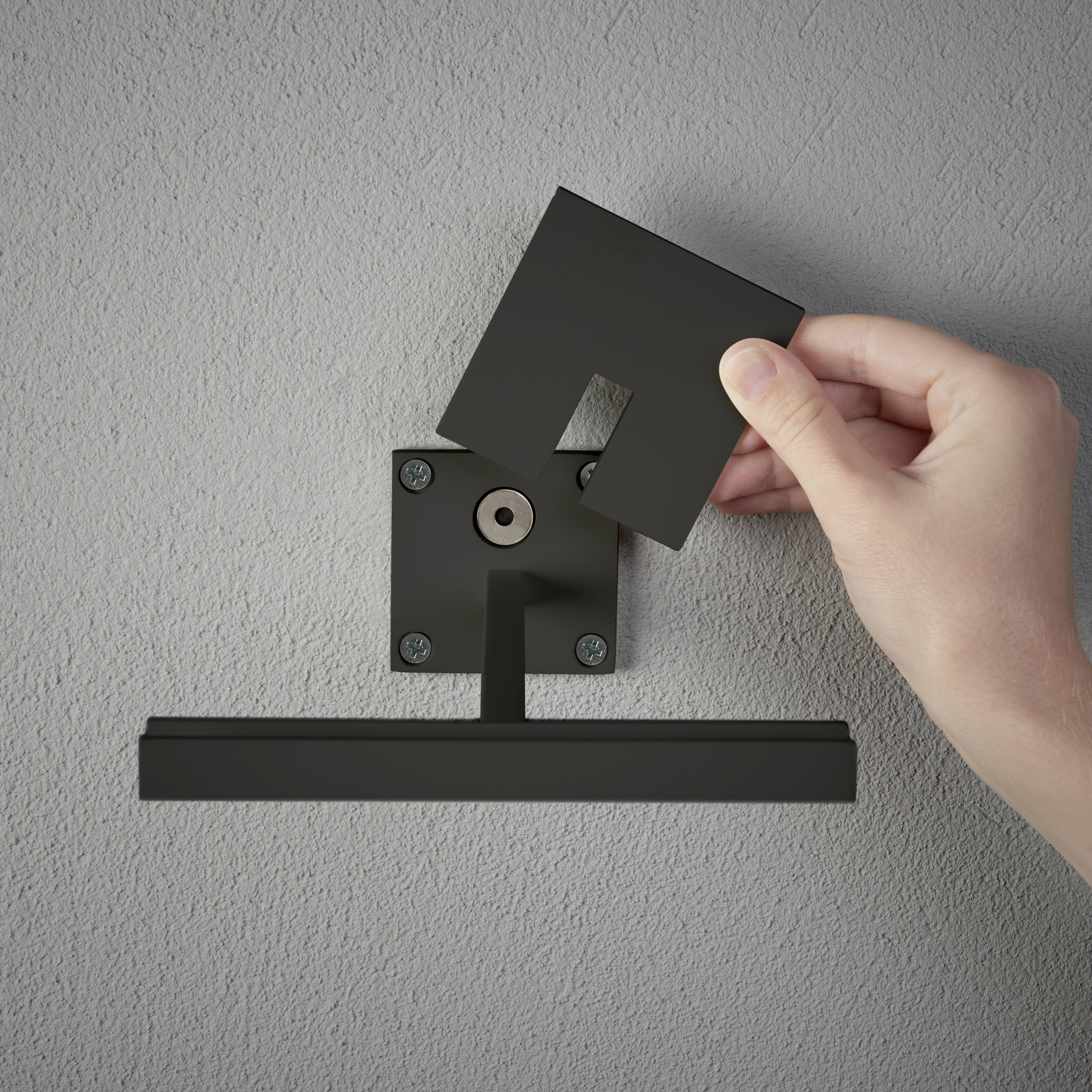 Une main tient un support noir rectangulaire contre le mur. Le support est composé de deux parties qui s'emboîtent l'une dans l'autre.