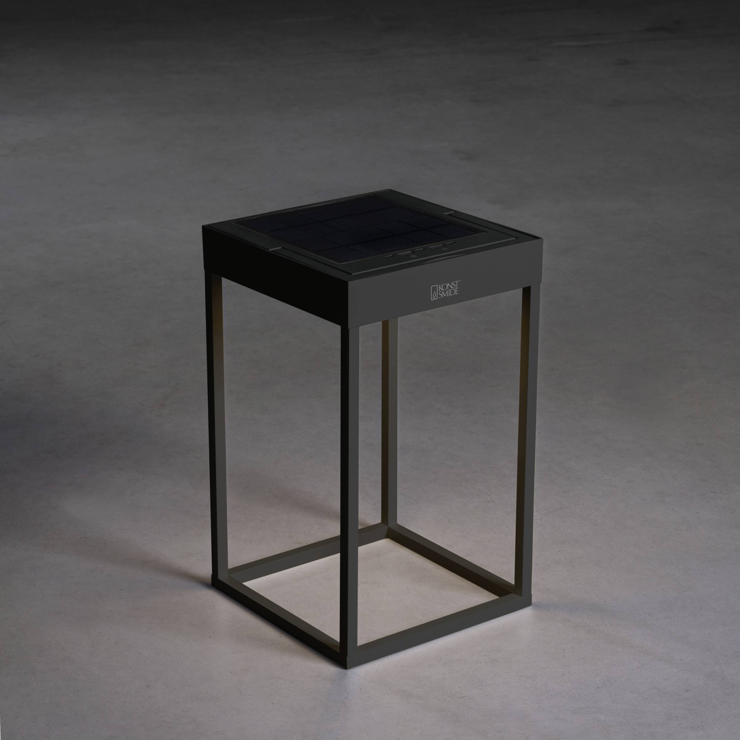 Une table d'appoint carrée noire avec un plateau en verre est posée sur un sol sombre.
