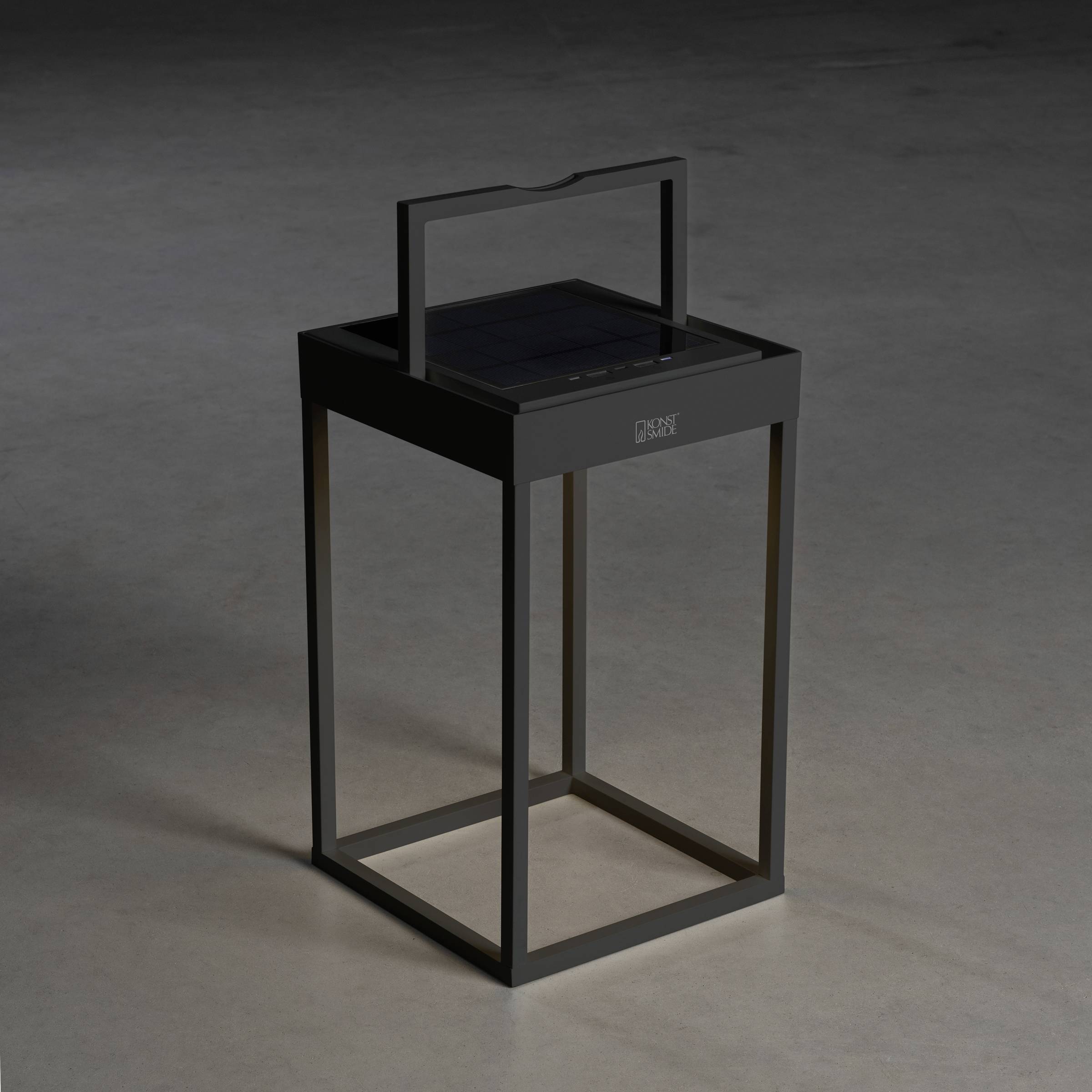 Une lanterne moderne noire avec un cadre rectangulaire est posée sur un sol gris. Elle présente des lignes épurées et une poignée en haut.