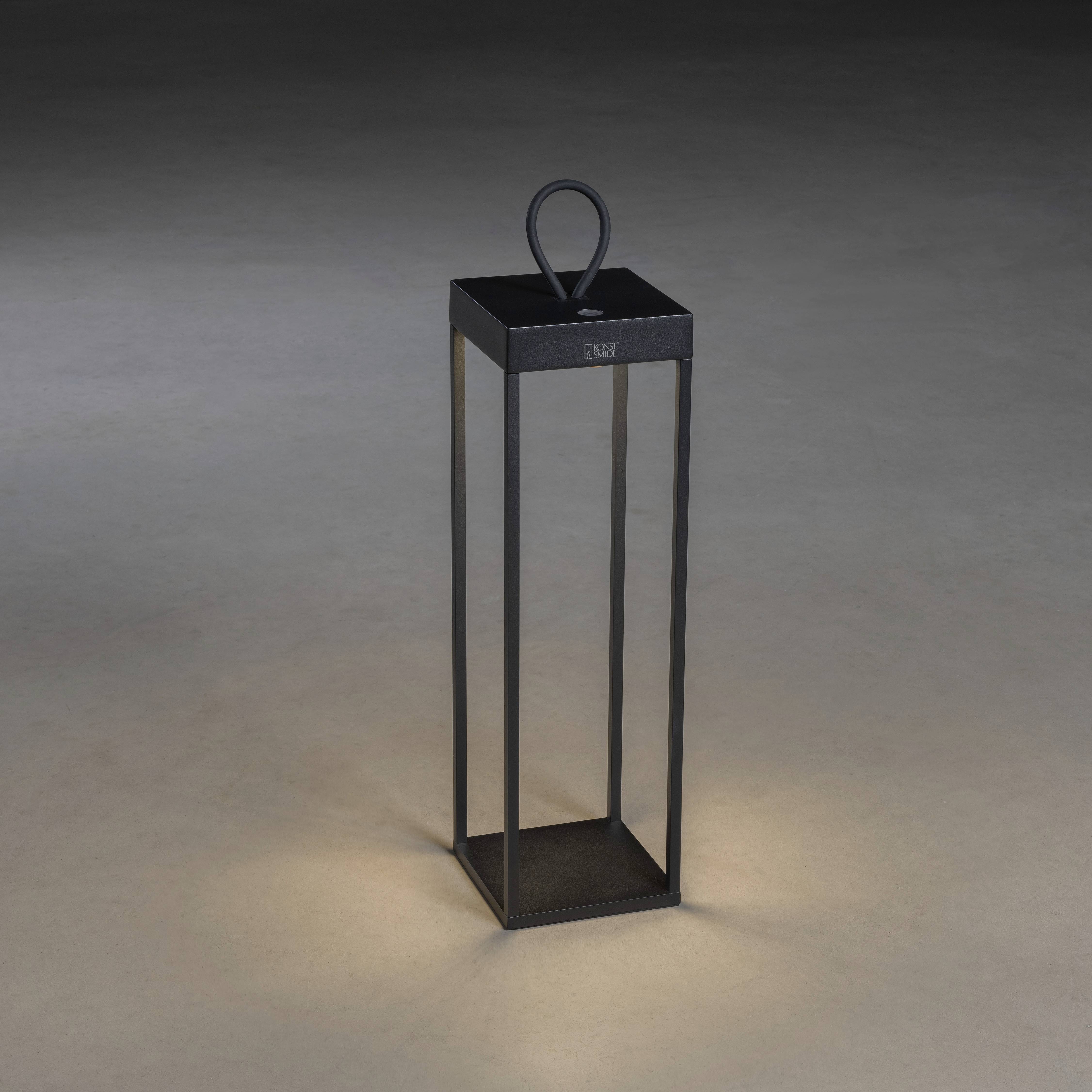 Une lanterne minimaliste noire aux parois vitrées repose sur un sol gris. Elle brille doucement et diffuse une lumière chaleureuse.