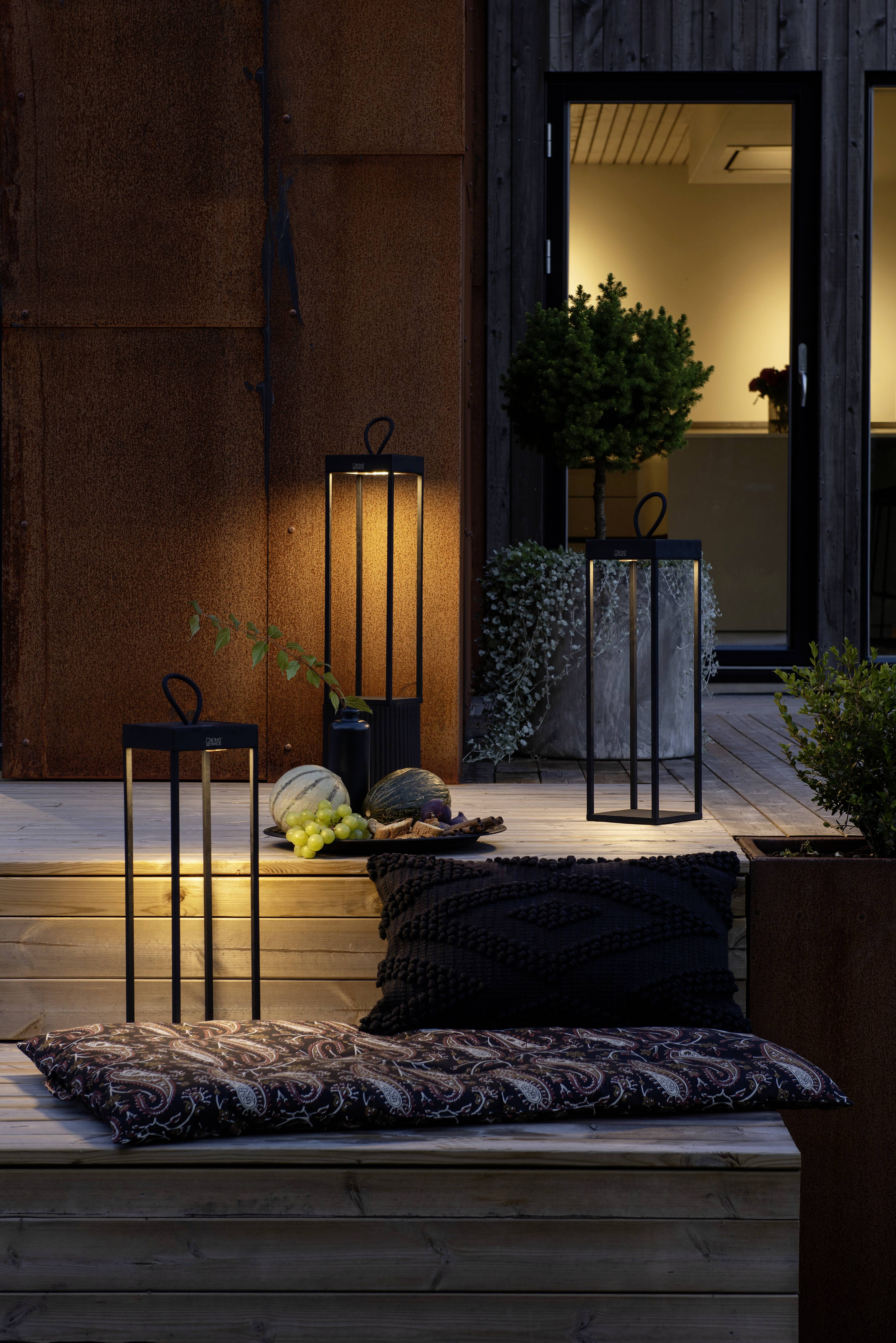 Des lanternes sur une terrasse en bois au crépuscule, avec des coussins au premier plan et une porte coulissante vitrée en arrière-plan.
