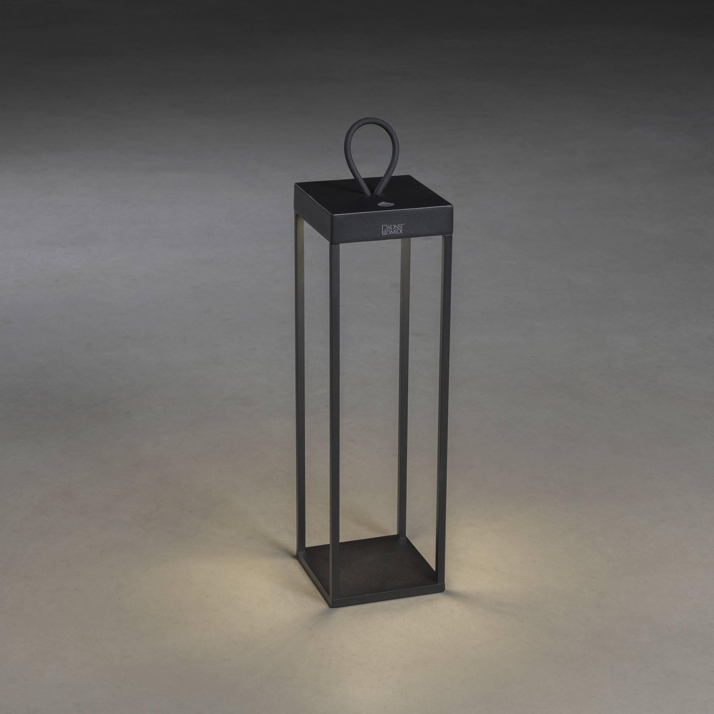 Lanterne noire, rectangulaire, avec une boucle de poignée, posée sur un sol gris. Émet une lumière douce vers le bas, à des fins décoratives.