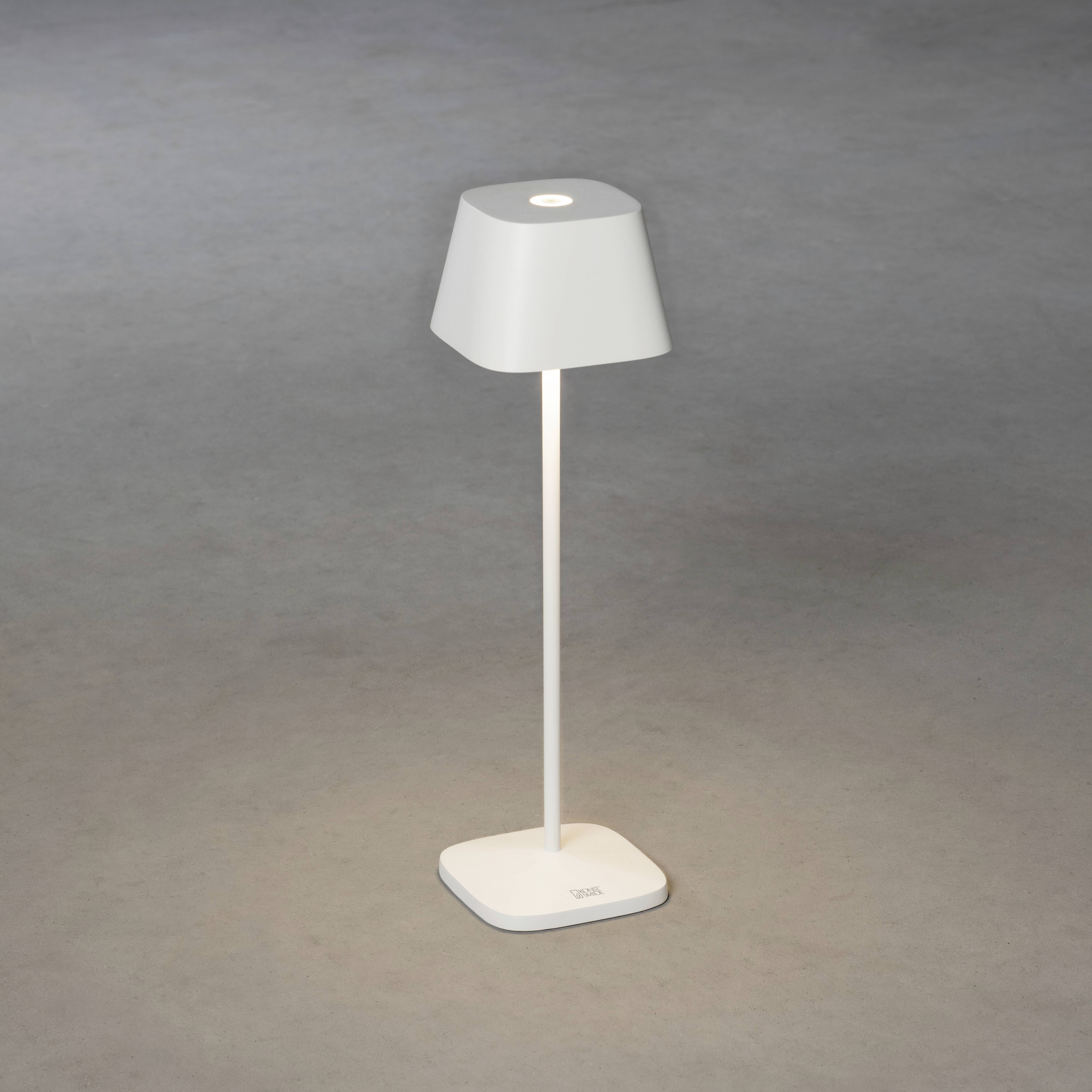 Lampe de table blanche minimaliste, avec un socle carré, posée sur un sol gris, dirigeant sa lumière vers le haut.