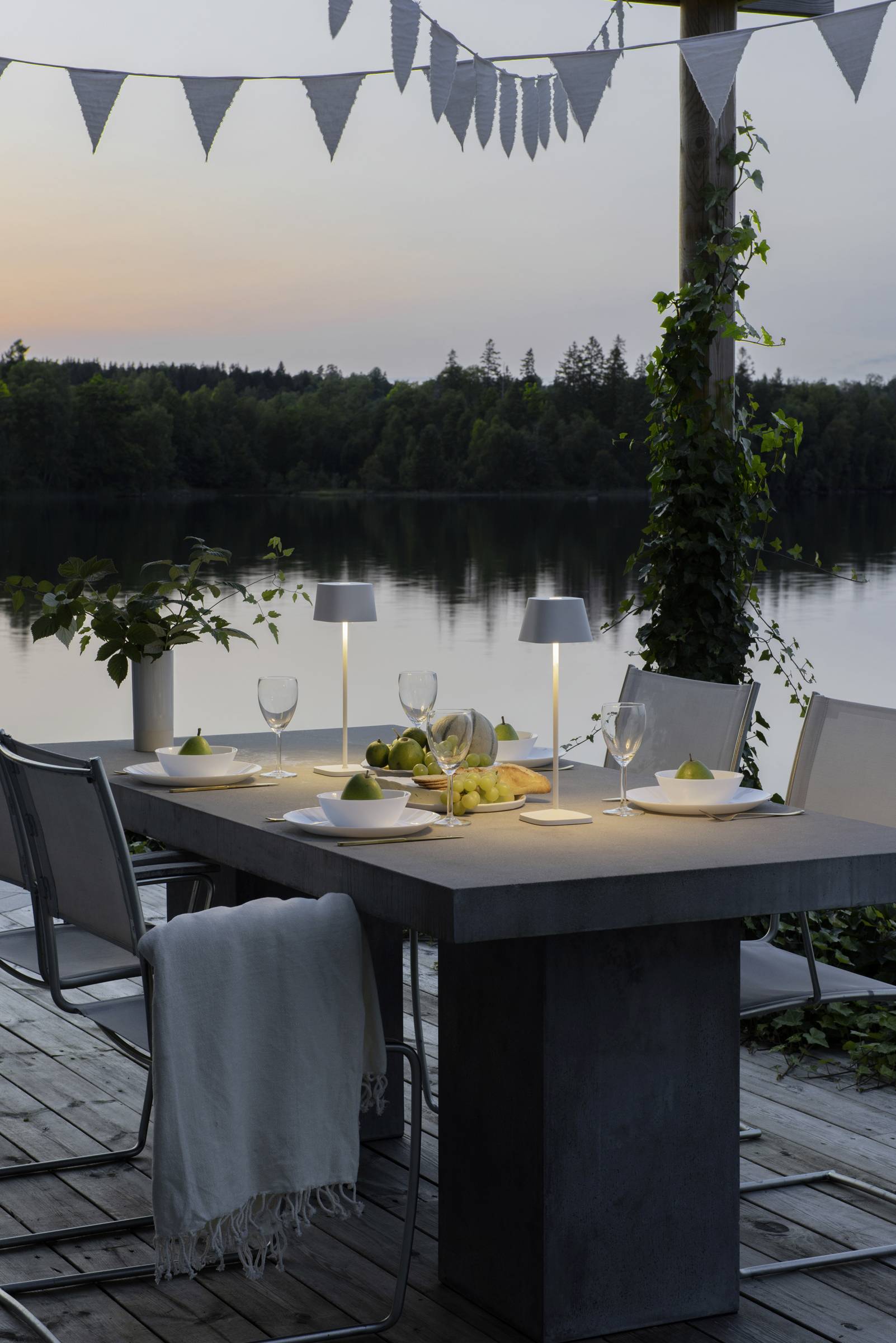 Une table de dîner en extérieur avec décoration et éclairage sur la rive du lac, entourée d'arbres et avec un coucher de soleil en arrière-plan.