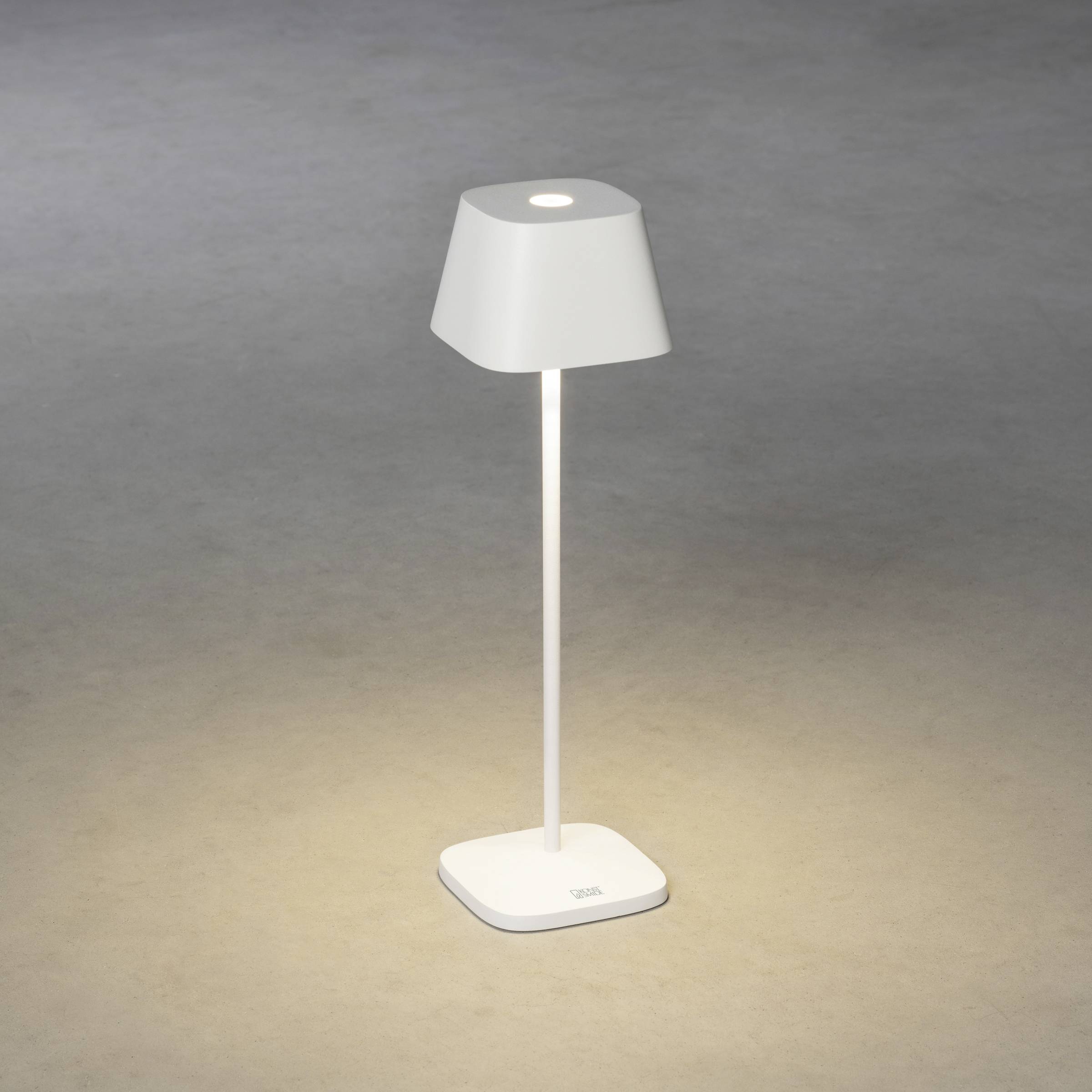 Une lampe de table moderne et minimaliste avec un abat-jour carré et un socle diffuse une lumière chaleureuse sur une surface grise.