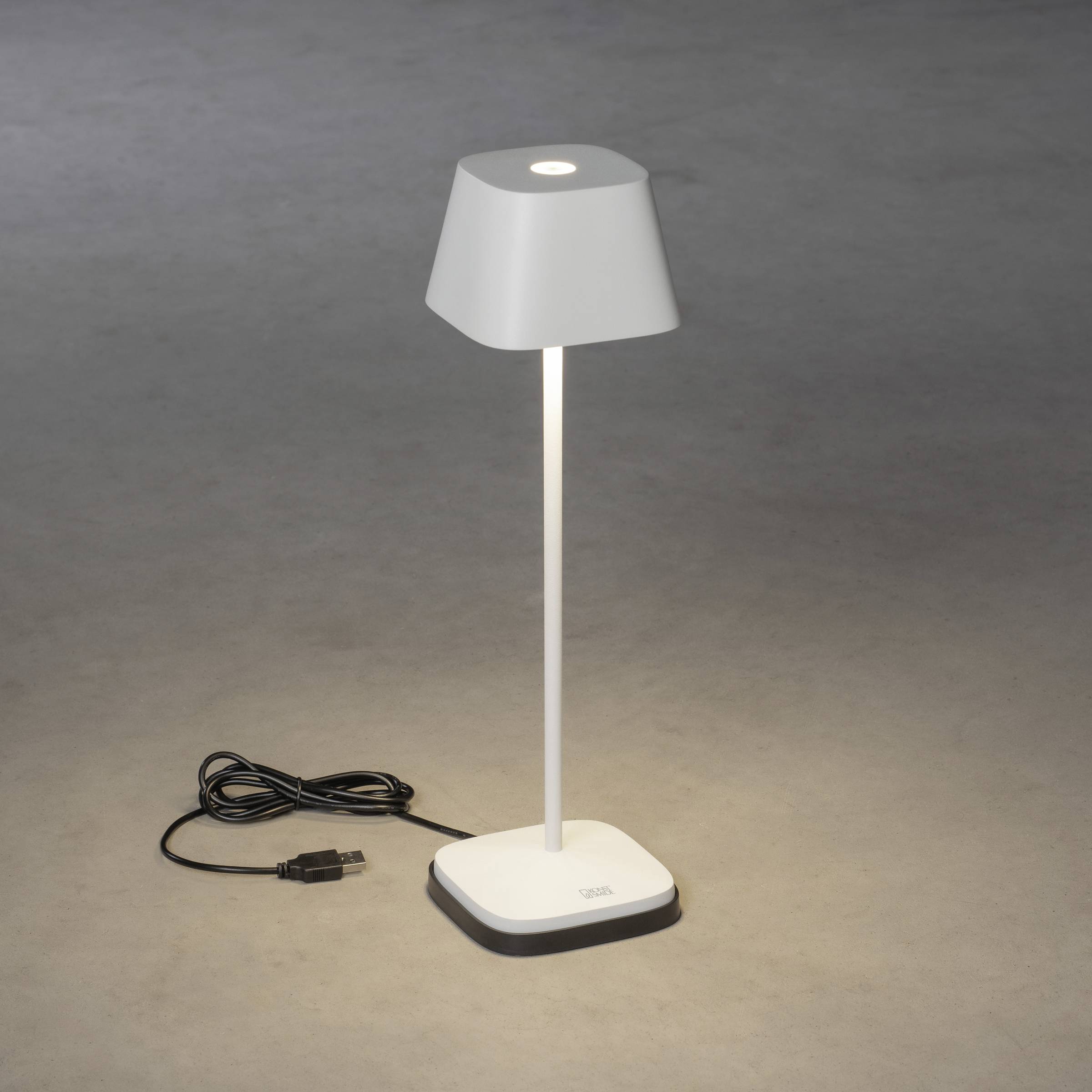 Une lampe de table blanche avec port USB sur un fond gris. La lampe est allumée et le câble est posé à côté.