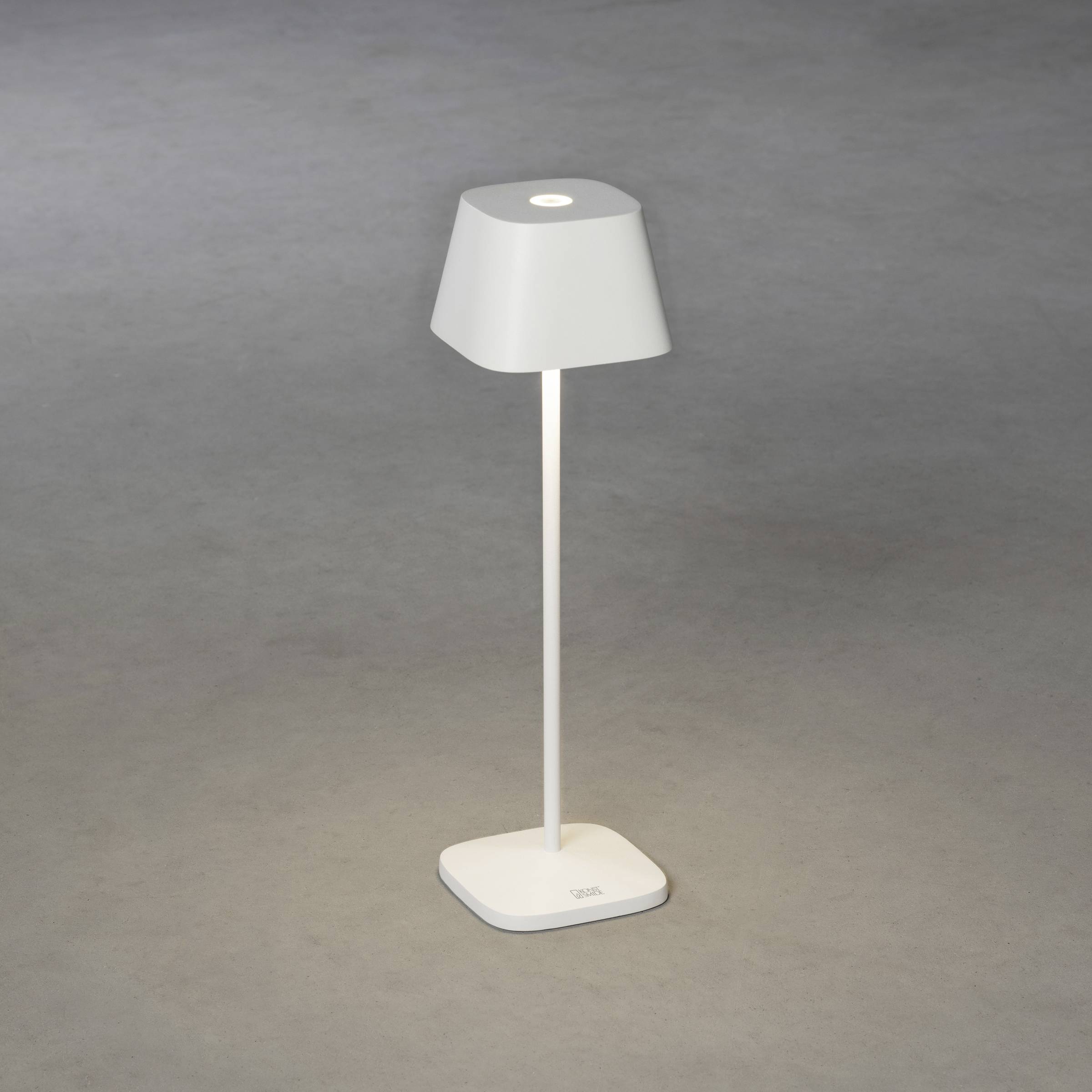 Une lampe de table moderne, blanche, avec un socle carré et un design épuré, est posée sur un sol gris.