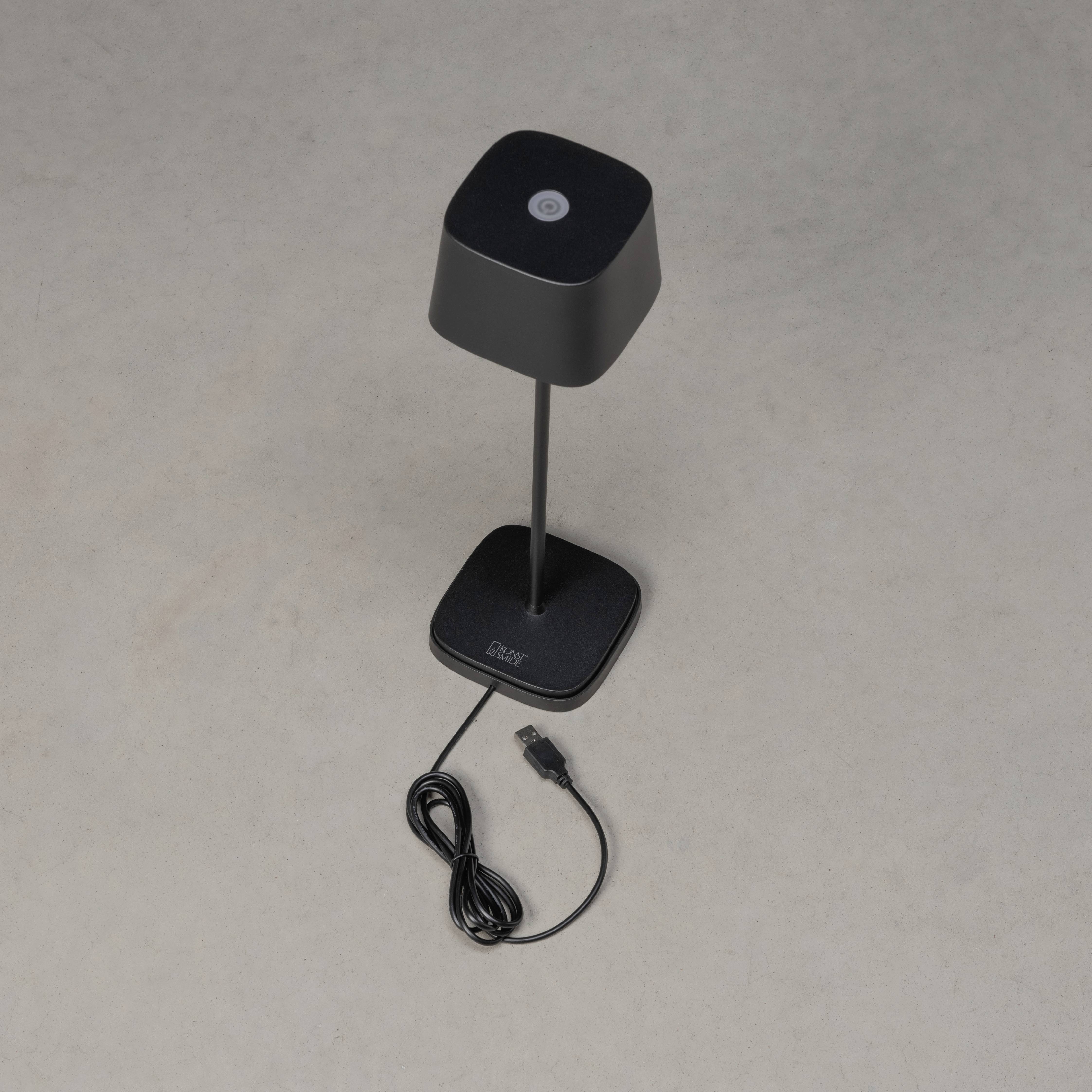 Une lampe de table noire avec un abat-jour carré et un câble de chargement USB sur un sol gris.