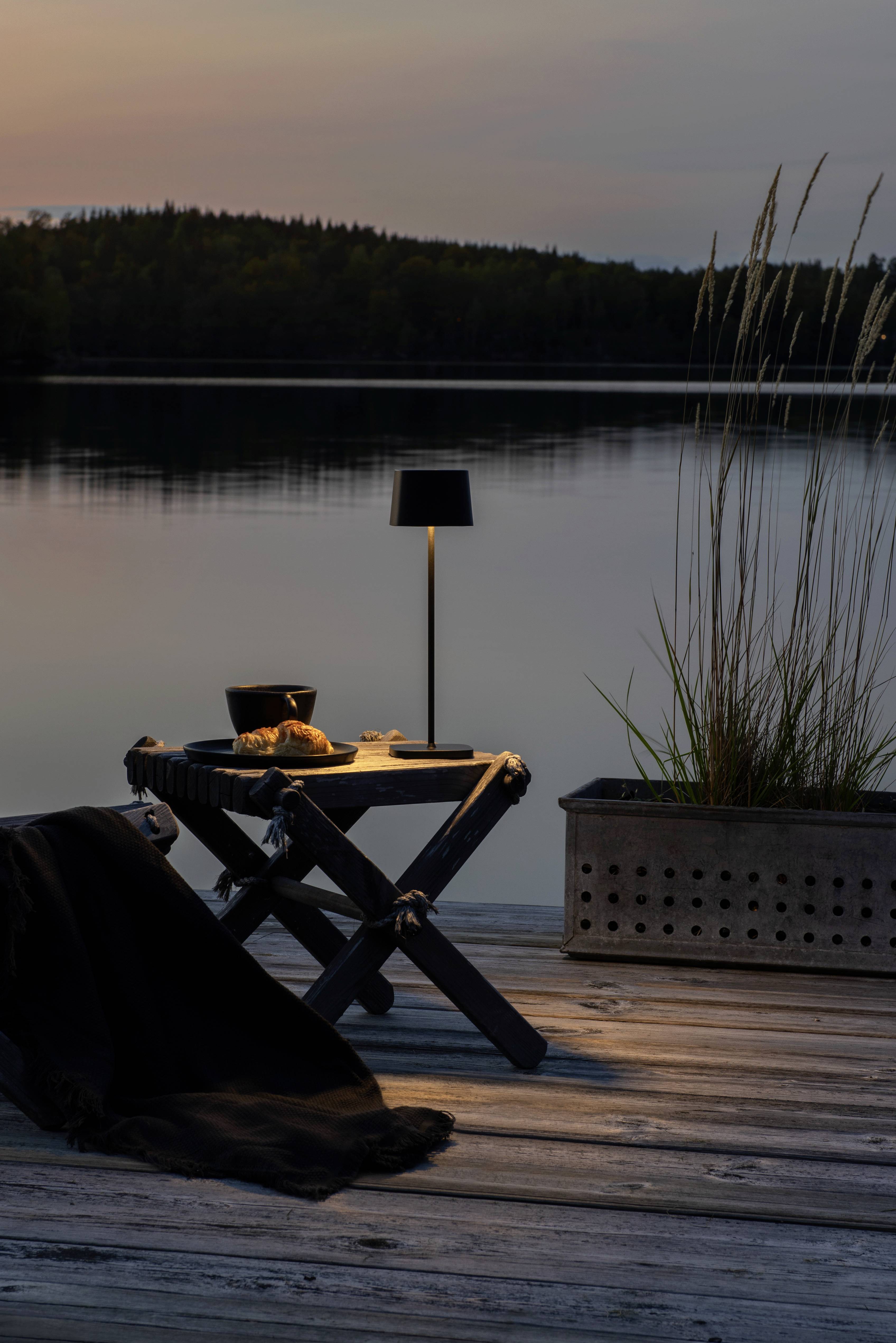 Une table en bois avec une lampe, un croissant et un tissu noir est placée sur une terrasse donnant sur un lac paisible au lever du soleil.