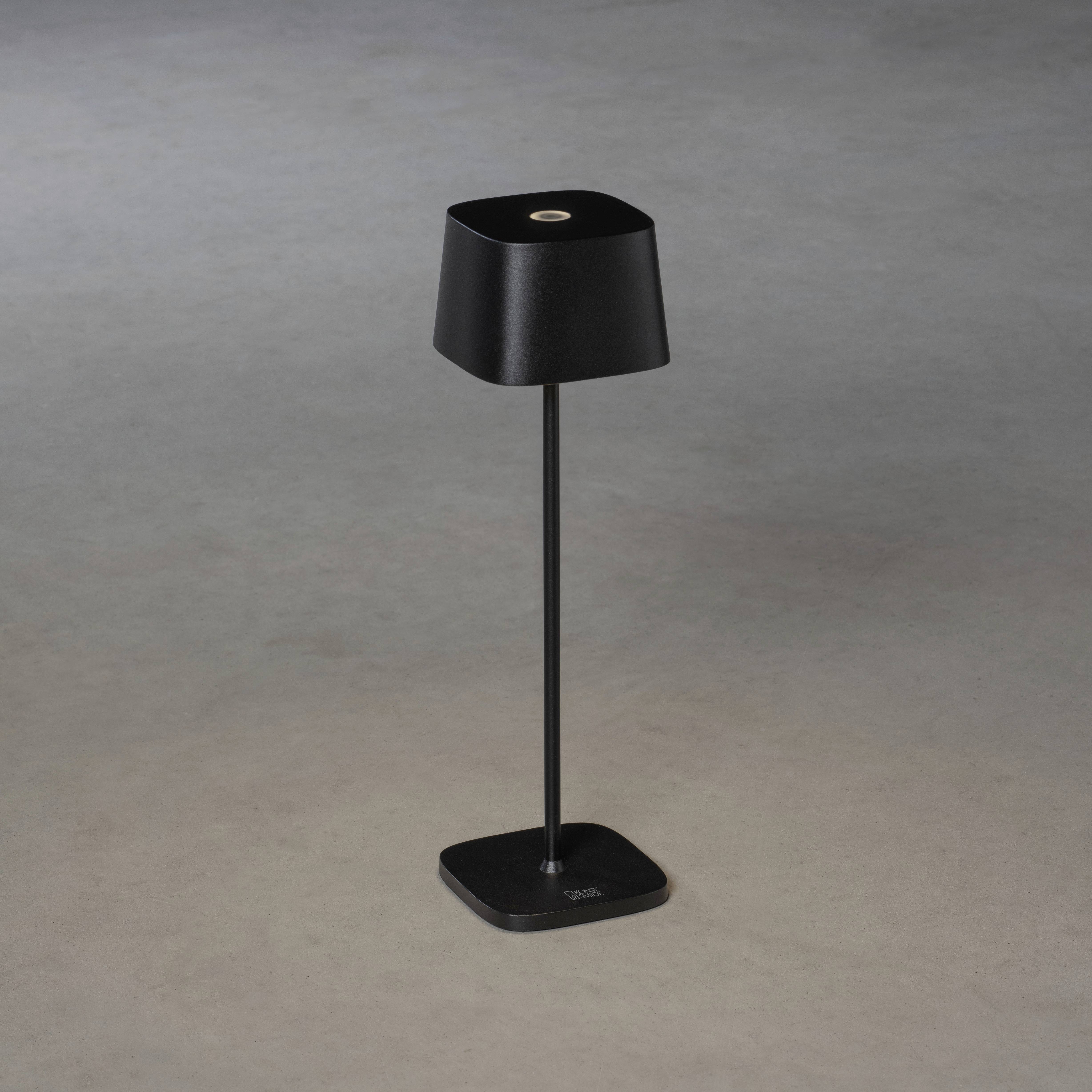 Une lampe de table noire, minimaliste, avec un abat-jour carré, reposant sur un support fin et stable, placée sur un fond gris.