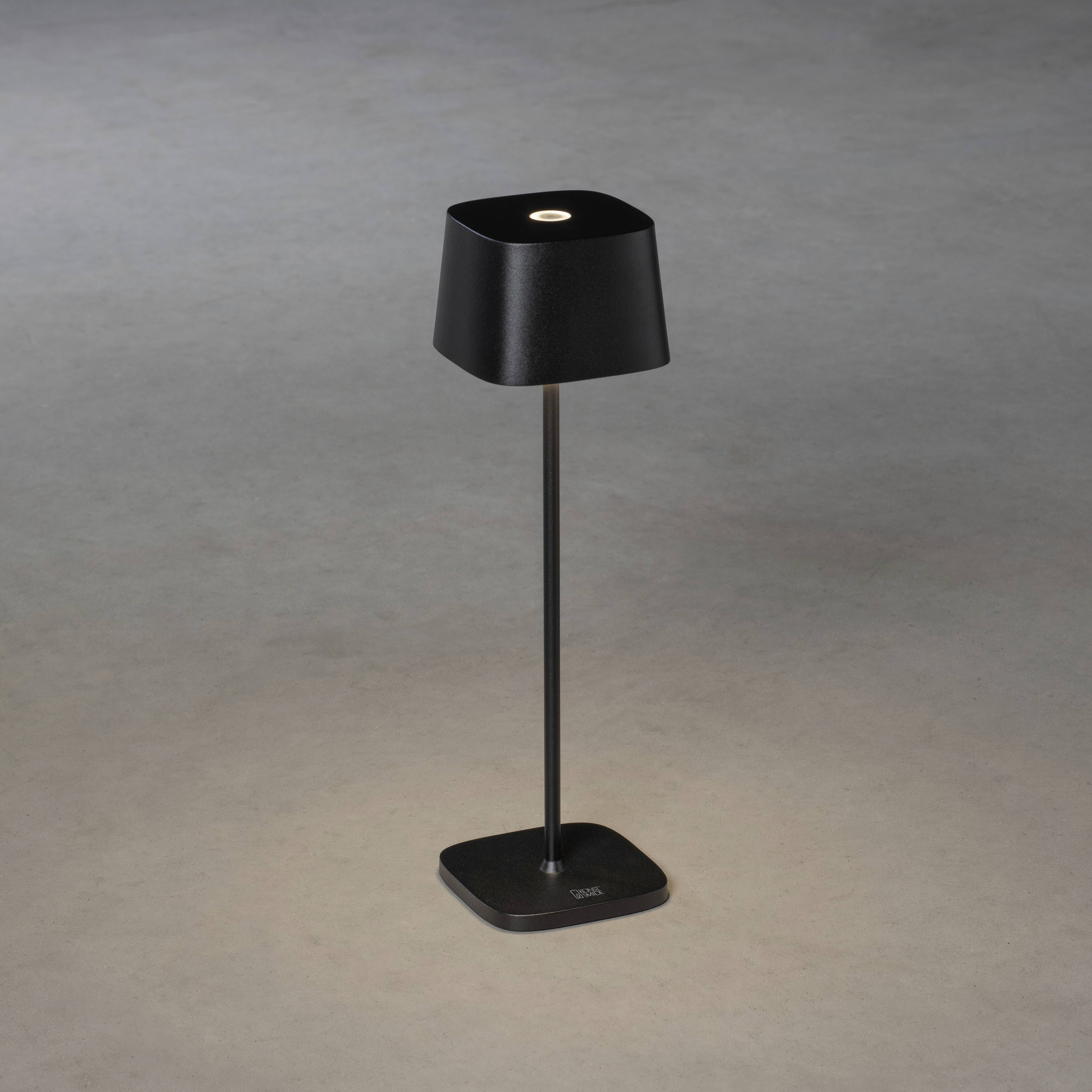 Lampe de table noire moderne avec un abat-jour carré, posée sur un sol gris, illuminant directement la zone située en dessous.