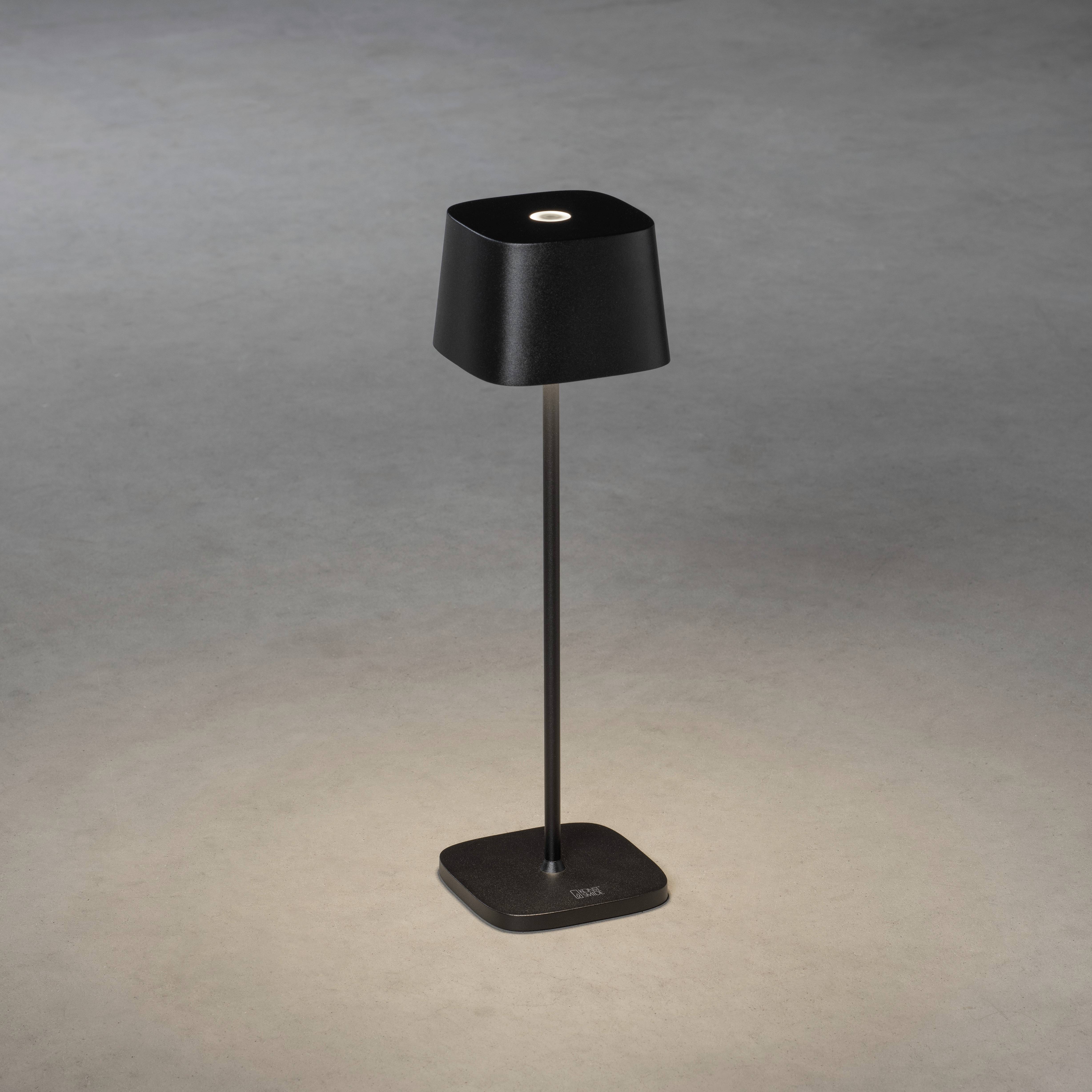 Une lampe de table moderne, noire, est posée sur un sol gris. La lampe présente un socle carré simple et épuré, et une tige élancée.