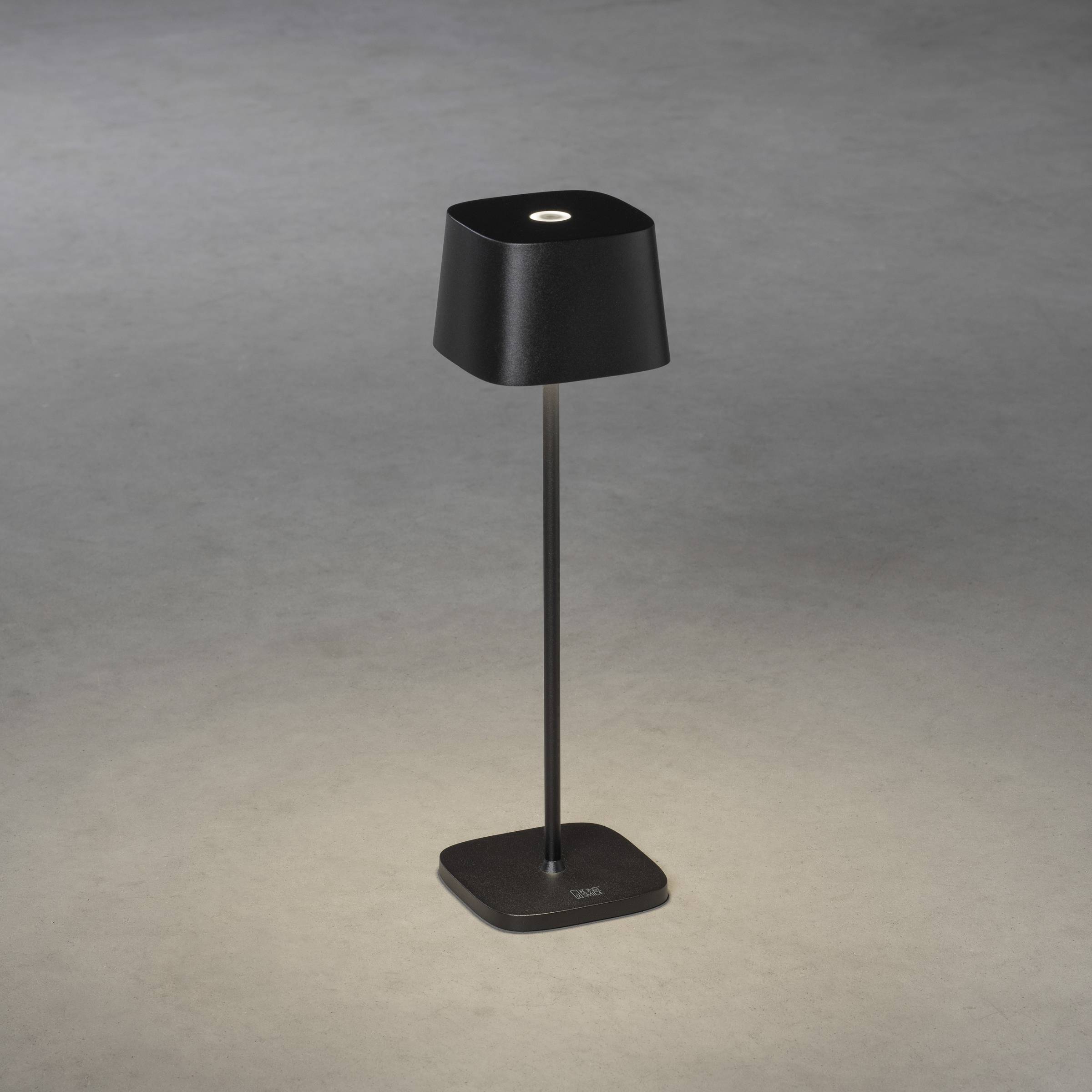 Lampe de table noire avec abat-jour carré sur fond gris. Lumière diffuse projetée vers le bas. Design moderne et minimaliste.