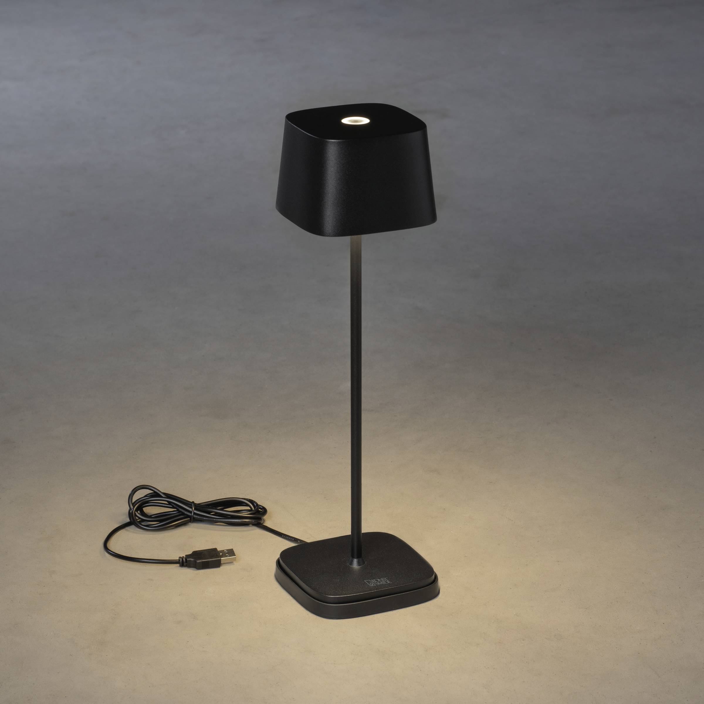 Lampe de table noire, sobre, avec connexion USB sur un fond gris. Elle éclaire vers le bas et illumine une petite surface.