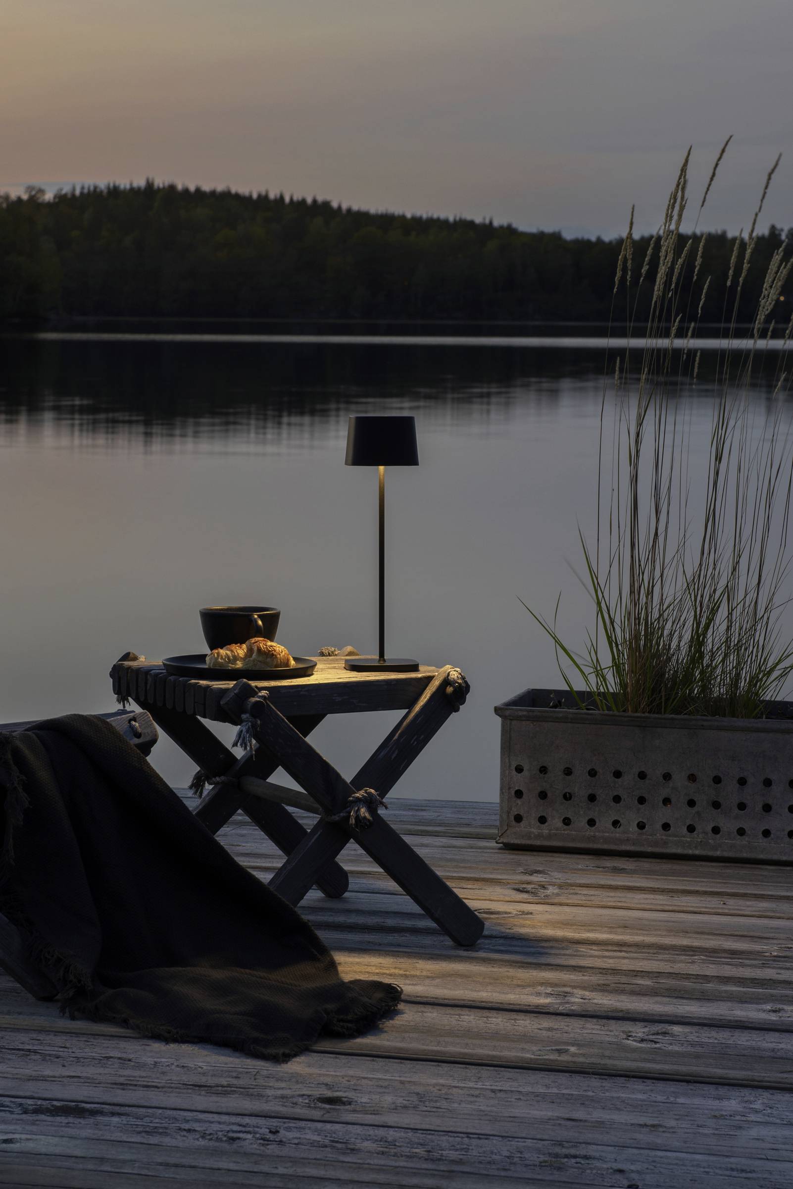 Un lac paisible au crépuscule, avec une table en bois, une couverture, une lampe et un pot de plantes sur la rive.