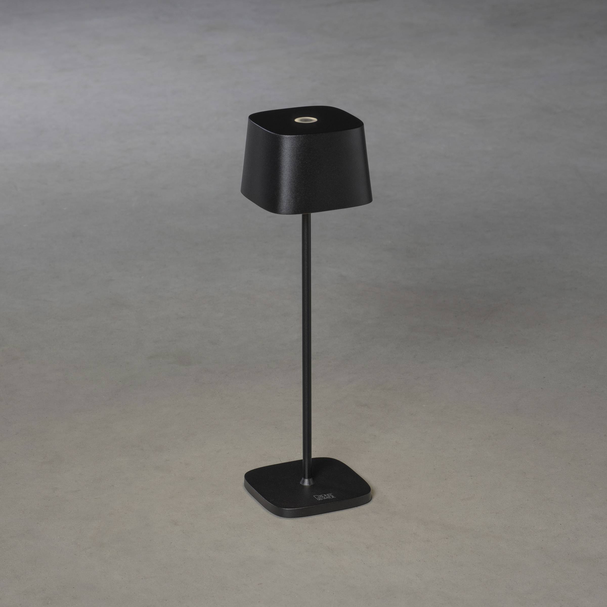 Lampe de table noire avec un abat-jour et un socle carrés, posée sur un sol gris. Design moderne et minimaliste.