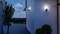 Un espace extérieur moderne au crépuscule, avec des appliques murales illuminées sur un bâtiment, des plantes en pots et une vue sur le paysage.