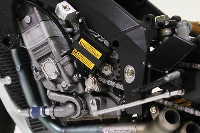 Gros plan d'un moteur de moto avec des détails visibles des chaînes, des tuyaux et des vis, partiellement recouvert d'autocollants.