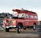 Un véhicule de pompiers rouge et ancien, équipé d'échelles et de sirènes, stationné sur un sol de gravier devant un tas de pierres.