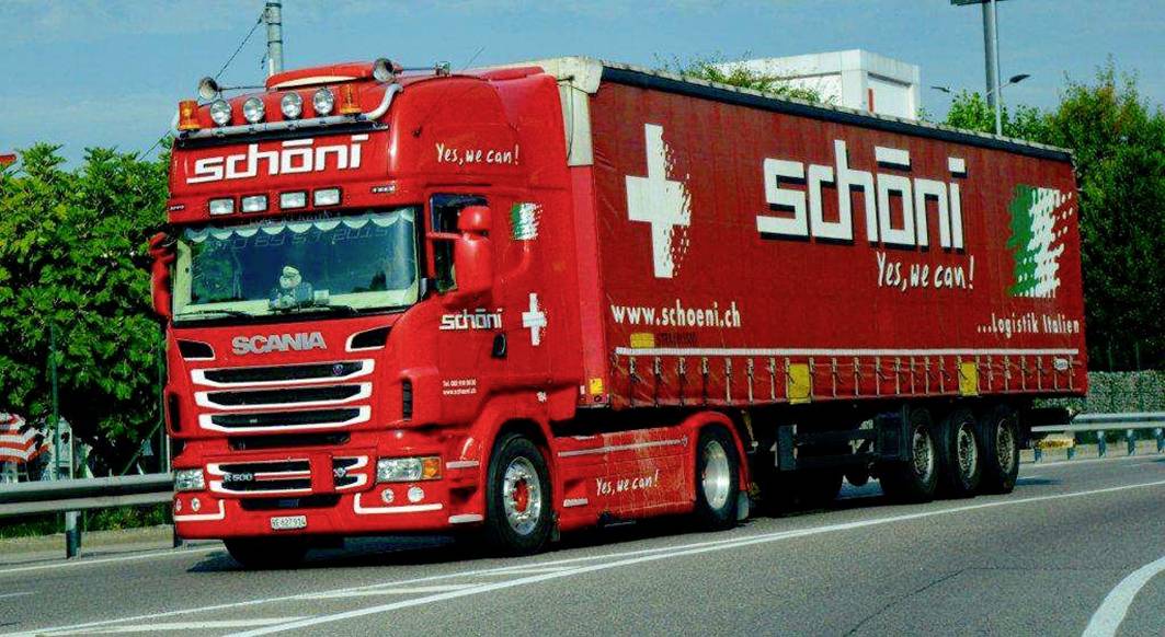 Un camion rouge portant l'inscription 'schöni' et 'Yes, we can!' circule sur une route. Le slogan et un drapeau suisse sont visibles.