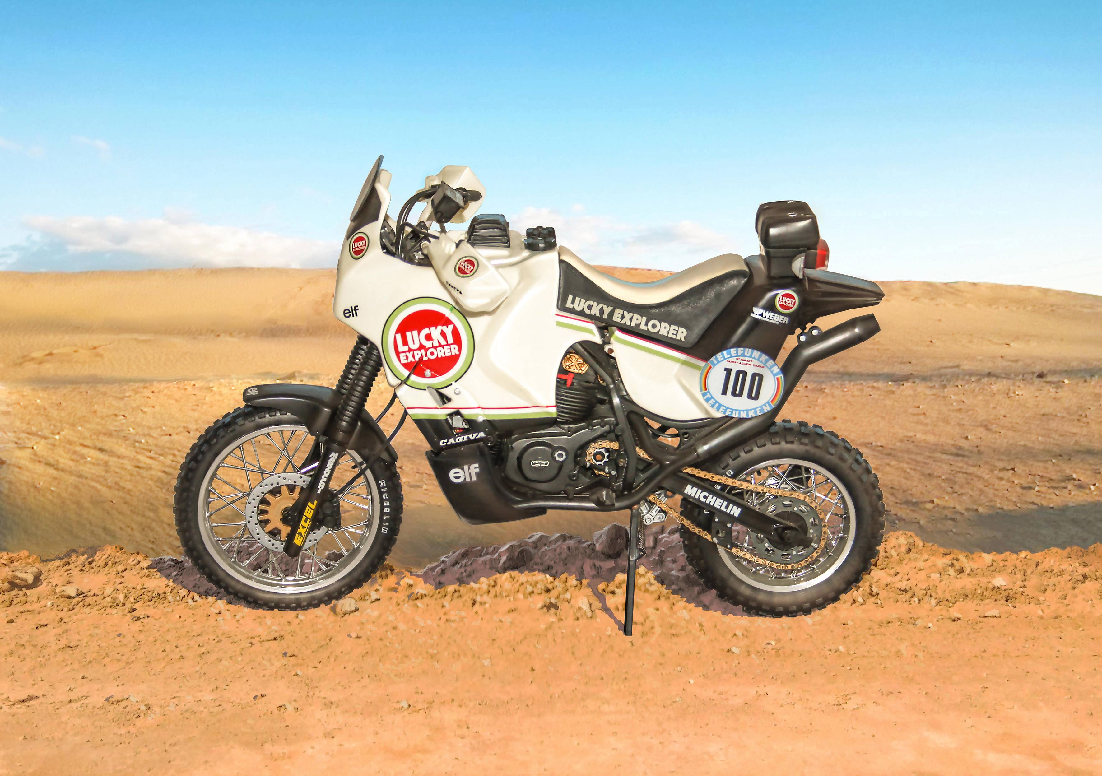 Une moto blanche portant l'inscription 'Lucky Explorer' est stationnée dans un paysage désertique. Elle arbore le numéro de course 100 et est équipée de pneus à gros profil.