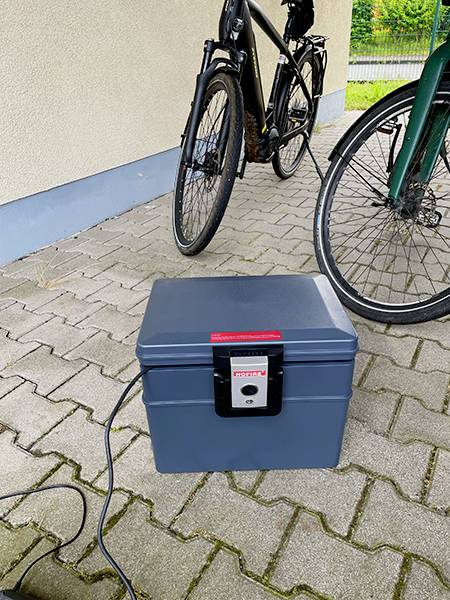 Boîte grise avec un câble posée sur le trottoir à côté de deux vélos.
