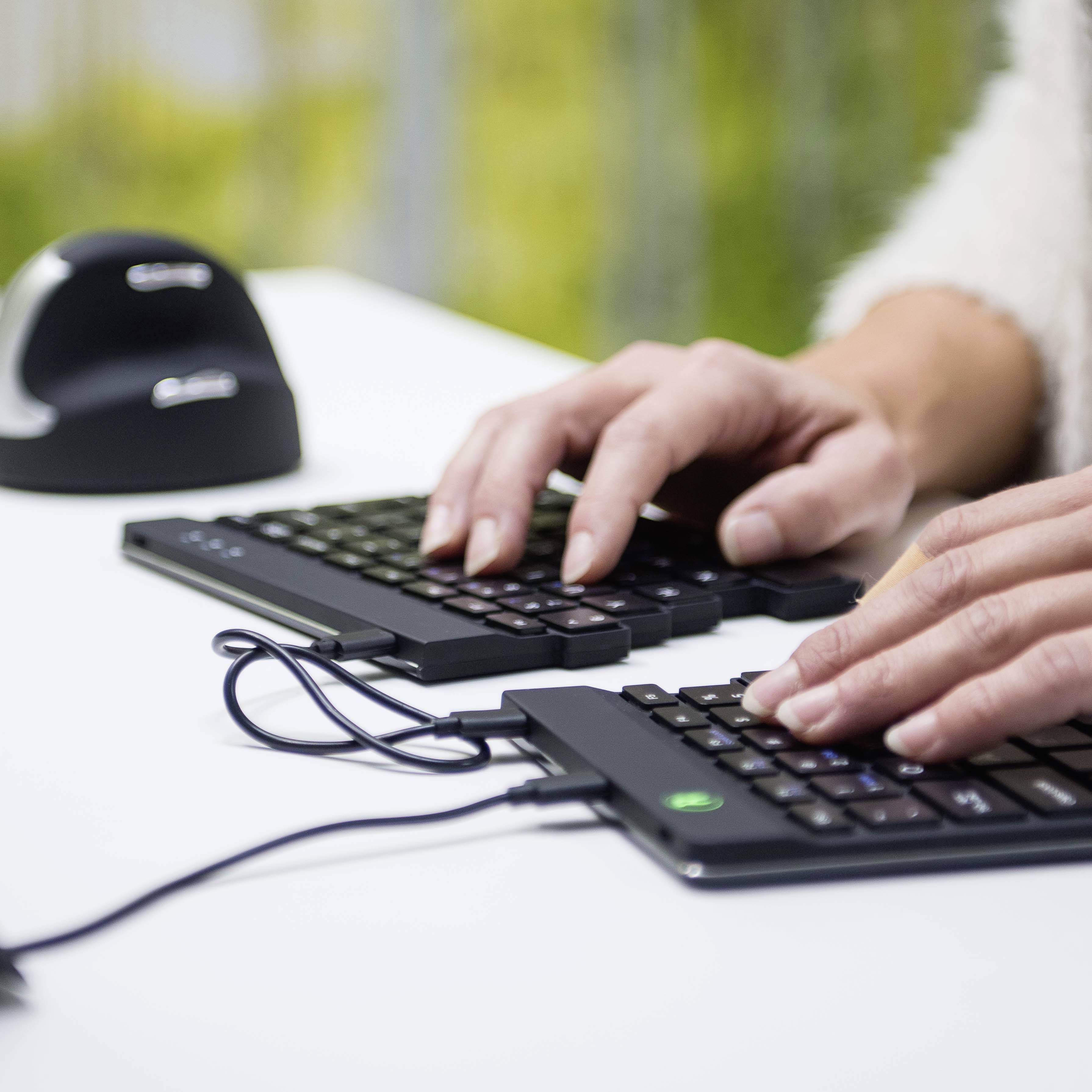 Une personne tape sur un clavier ergonomique divisé, posé sur une table. Un arrière-plan naturel flou est visible.