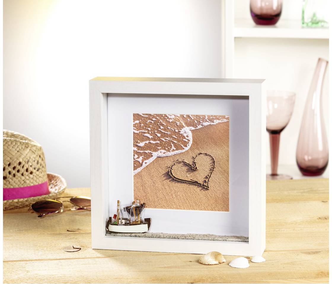 Un cadre avec un cœur dessiné dans le sable devant une vague. Au premier plan, un chapeau, des lunettes de soleil et des coquillages sont posés sur une table.