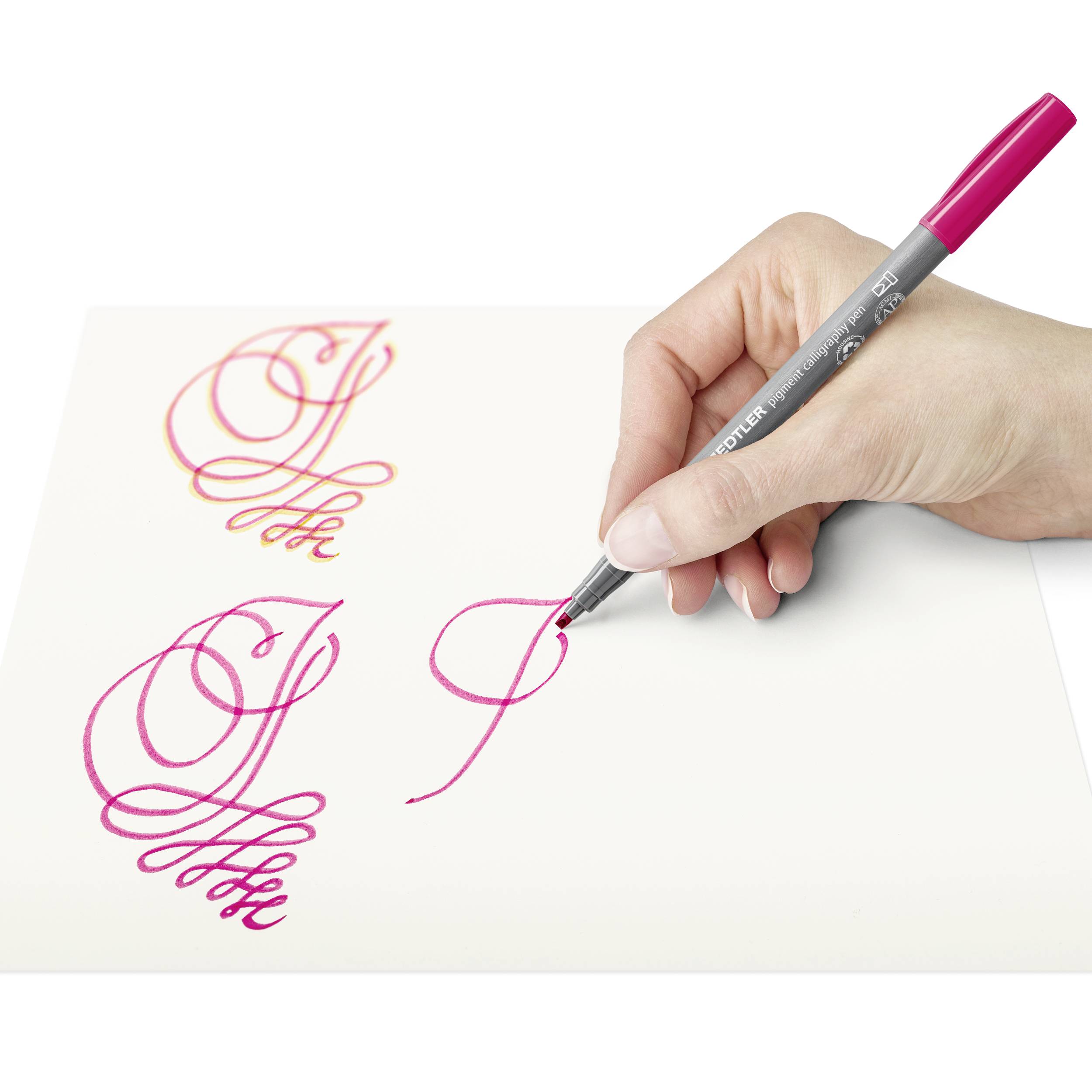 Une main écrit des lettres calligraphiques avec un stylo rose sur du papier blanc.