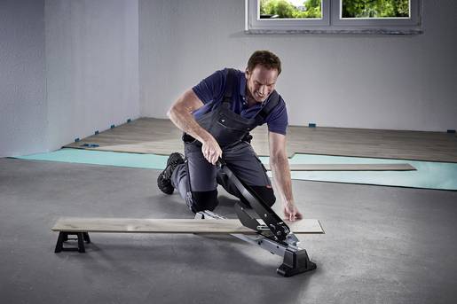 Un homme en vêtements de travail coupe une planche de bois avec un outil sur le sol d'une pièce inachevée.