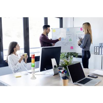 Deux personnes dans un bureau : deux sont debout devant un tableau blanc, discutant des graphiques et des notes ; une est assise à la table avec une tasse de café.