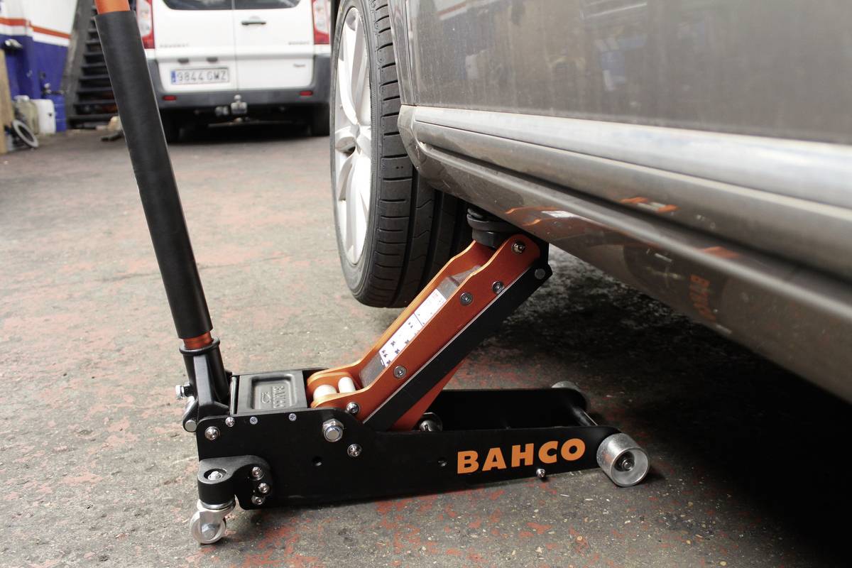 'Un cric soulève une voiture au niveau du bas de caisse. Dispositif de levage avec levier et roues sur le sol d'un atelier, roues du véhicule partiellement visibles.'