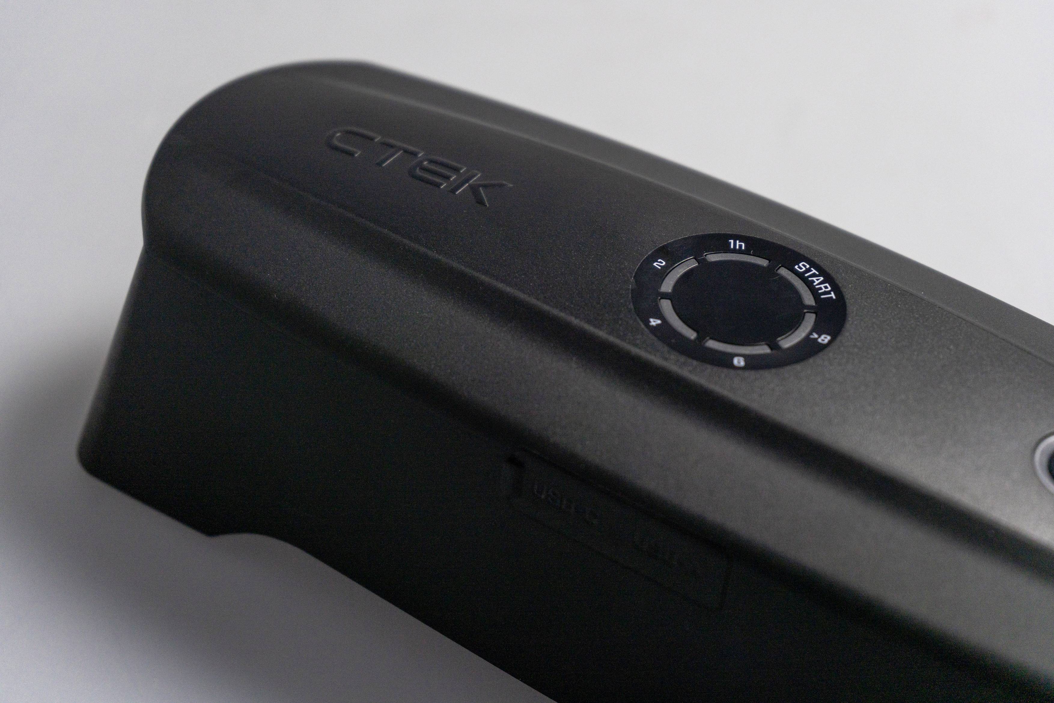 Un chargeur noir portant l'inscription 'CTEK' avec un panneau de commande circulaire présentant des réglages de temps, posé sur une surface grise.
