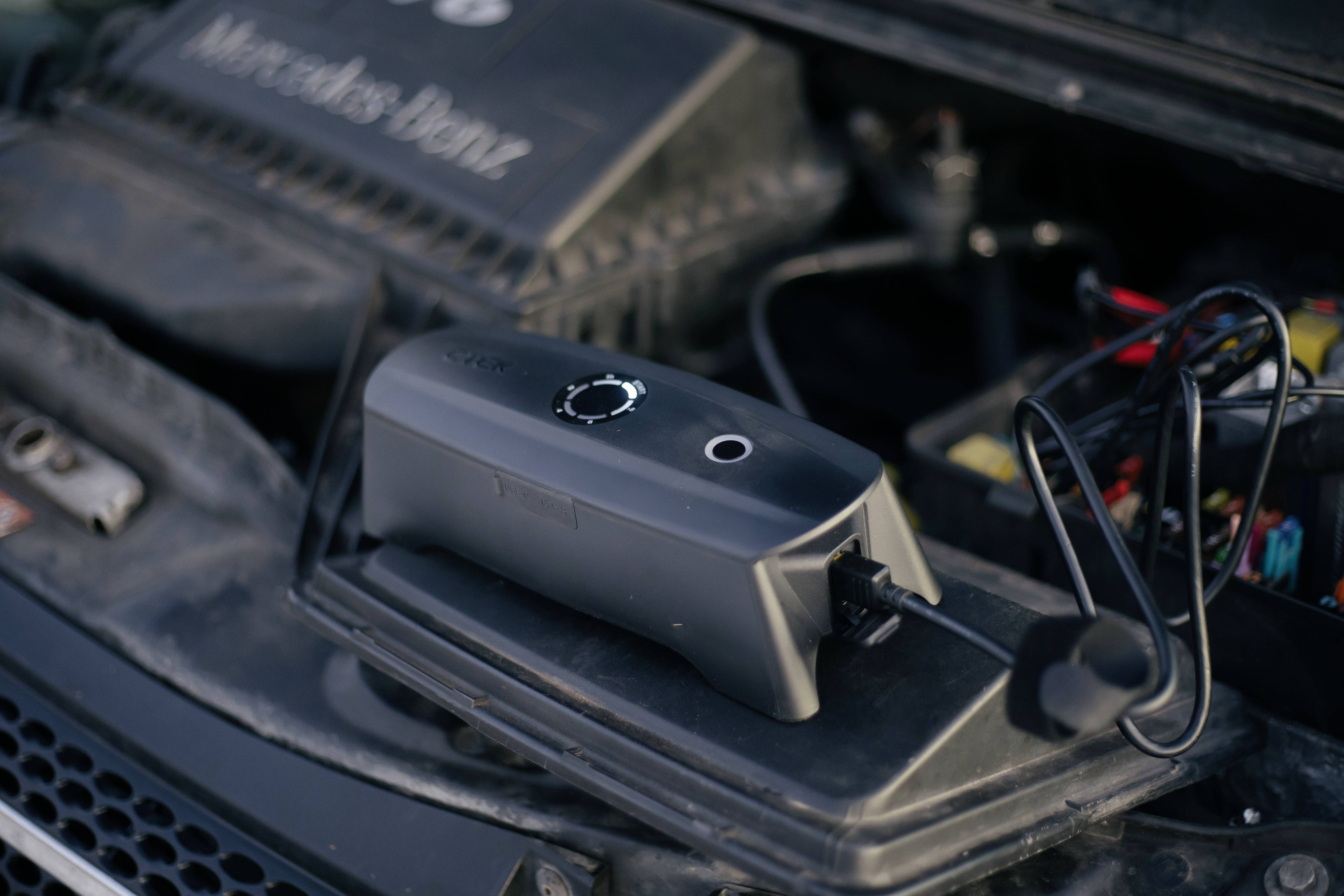 Une voiture est en train d'être rechargée avec un chargeur mobile placé sur le moteur. Des câbles sont visibles, le capot du moteur est ouvert.